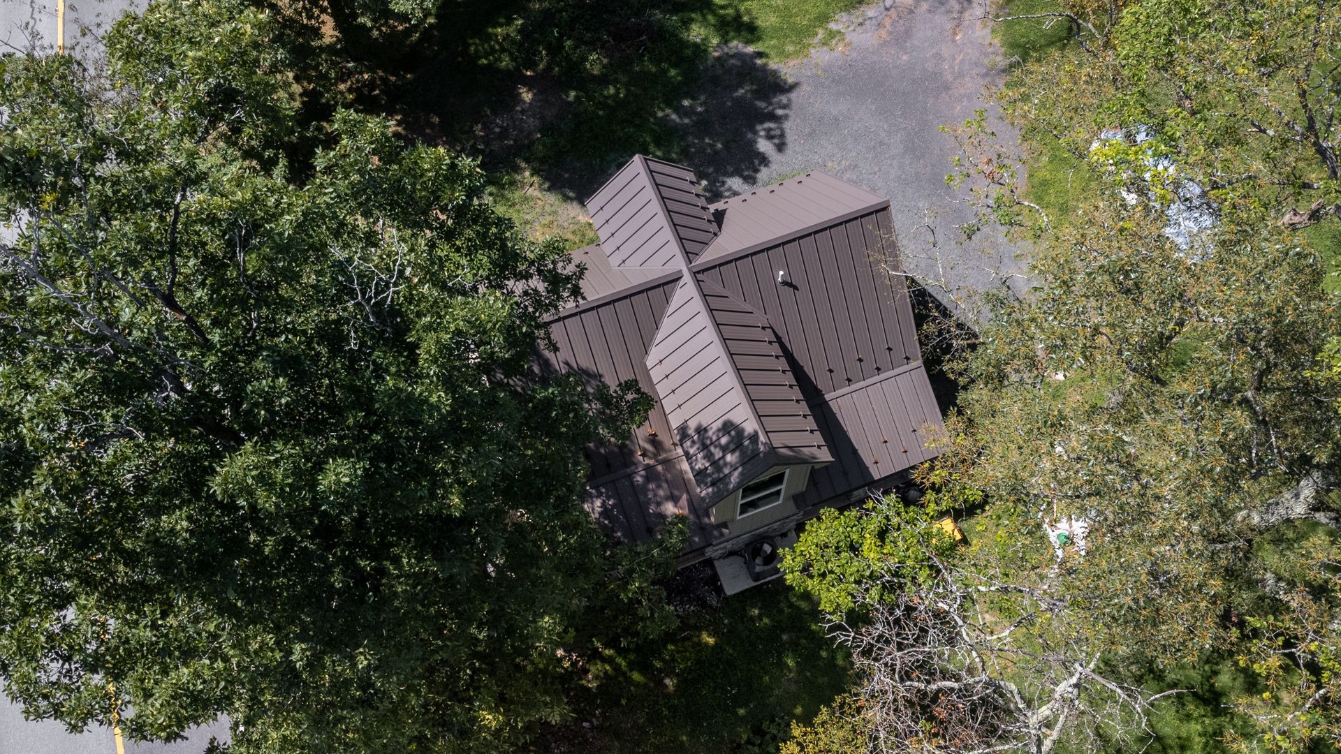 Overhead view of a brown-roofed house surrounded by green trees, gravel driveway, and road.