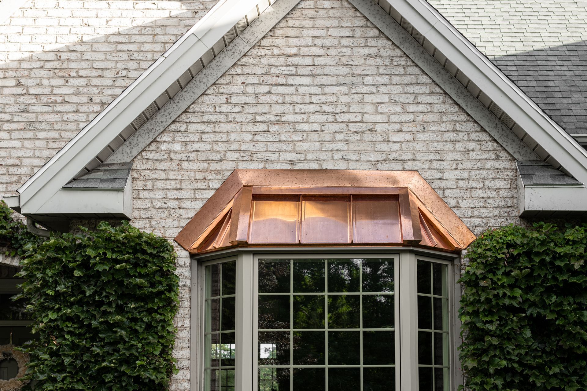 Bay window with copper roof, set in white brick wall, framed by green bushes.