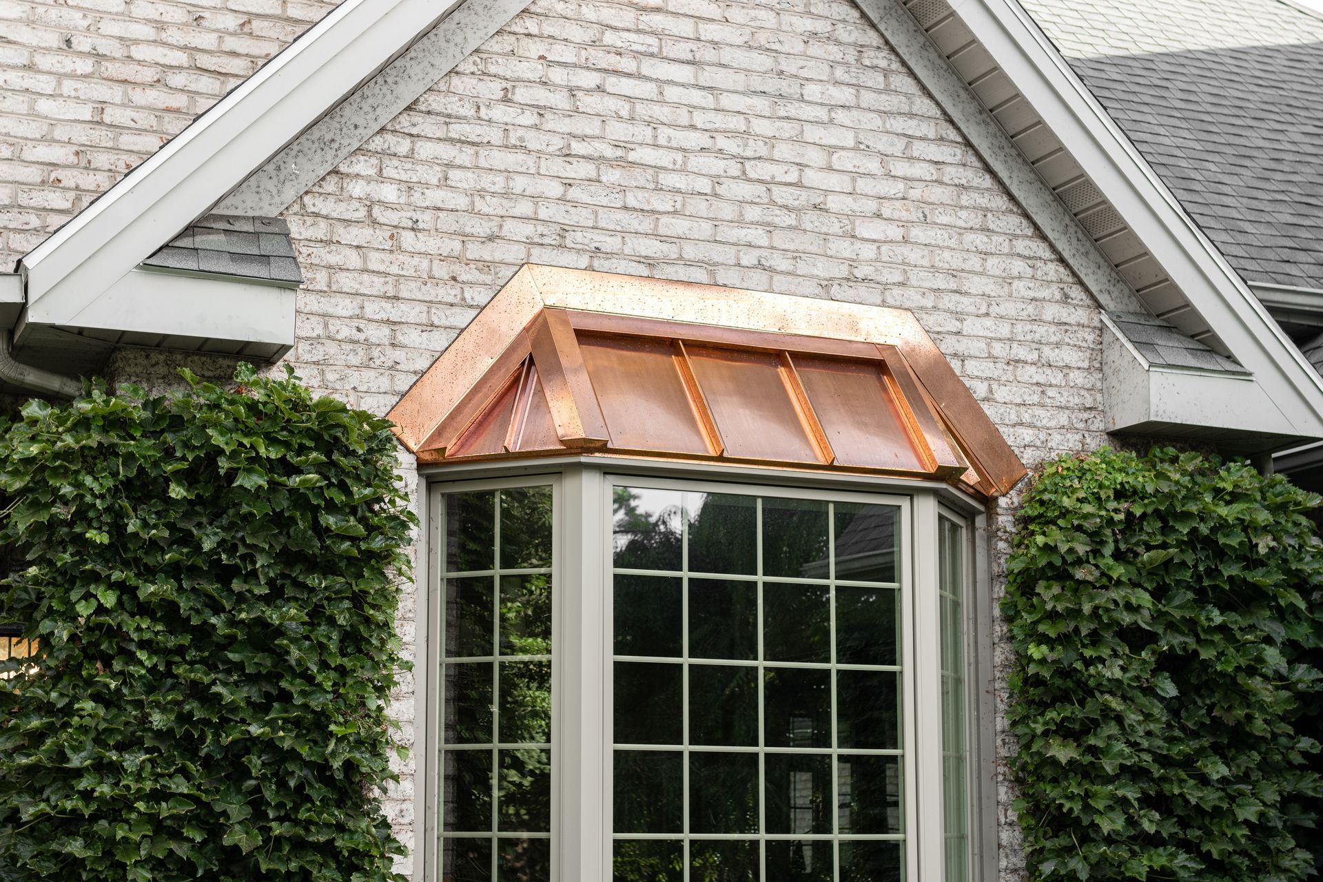 Copper-roofed bay window on a brick house. Ivy bushes flank the window.