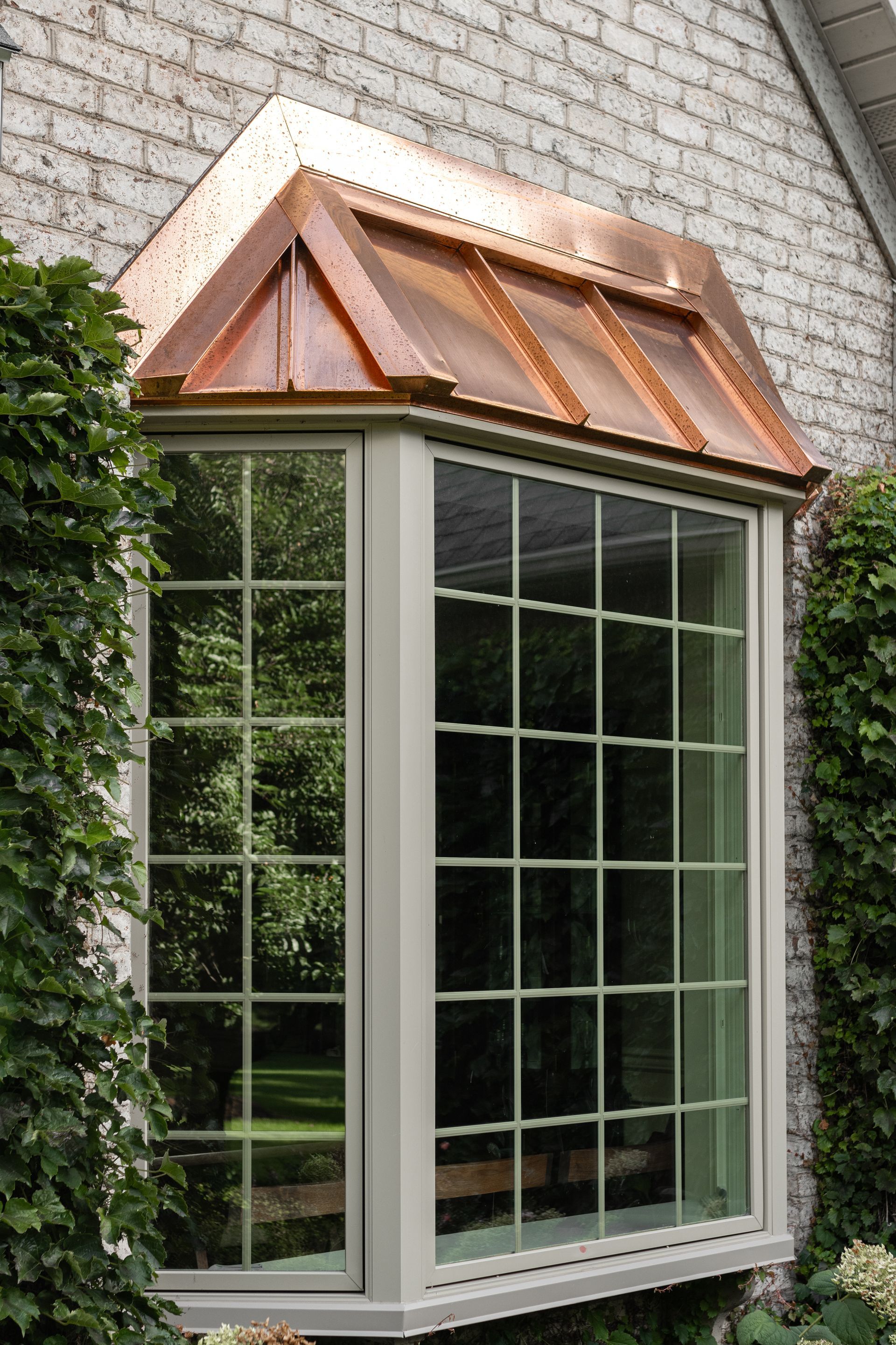 Bay window with copper roof, cream trim, and multi-pane windows against a brick wall.
