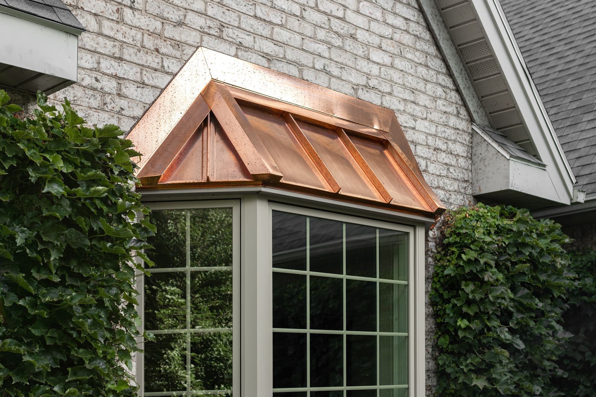 Copper-roofed bay window with a shingle siding exterior. Lush green bushes flank the window.