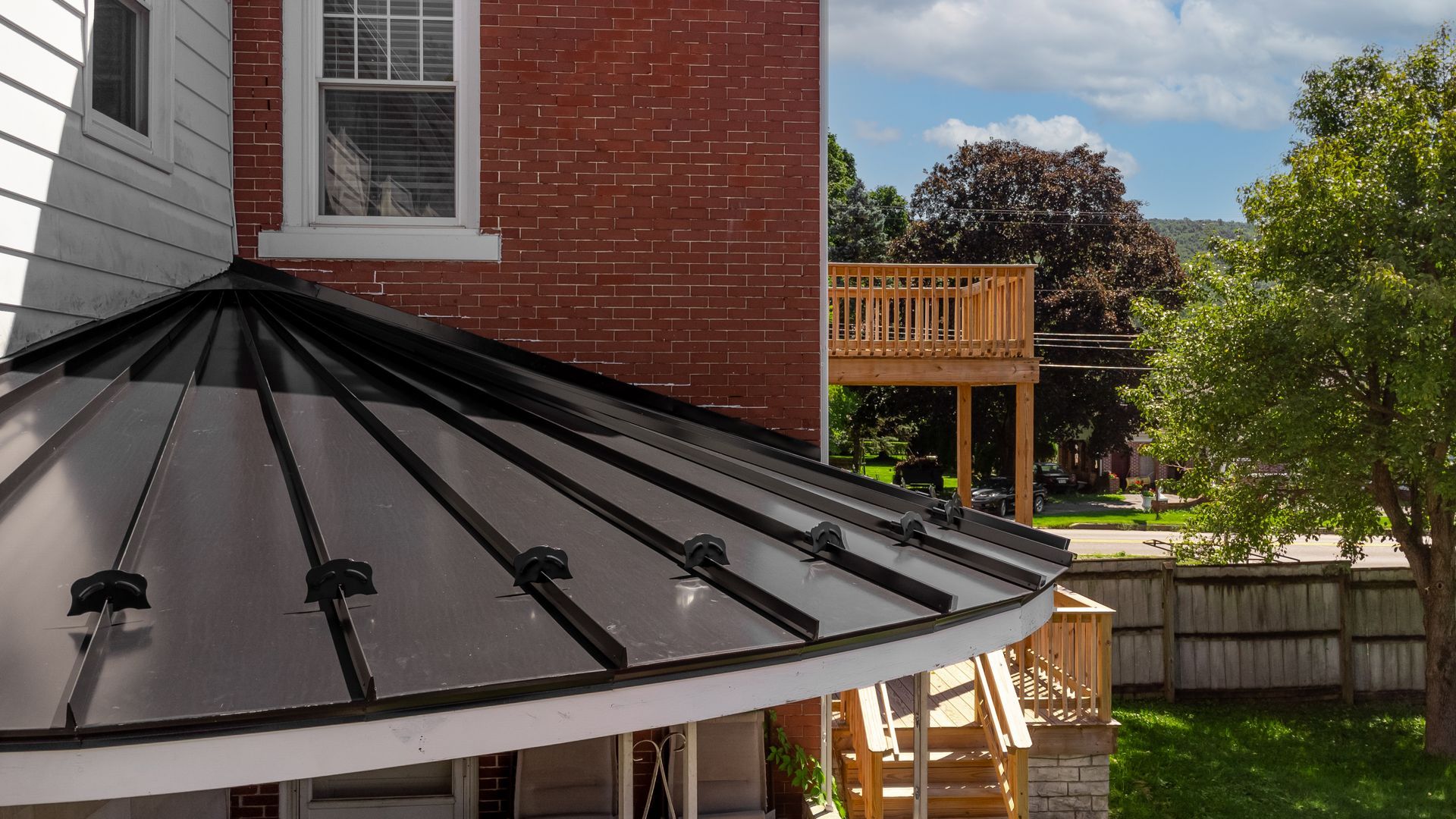 A black metal roof over a curved porch, attached to a red brick building with a wooden deck visible in the background.