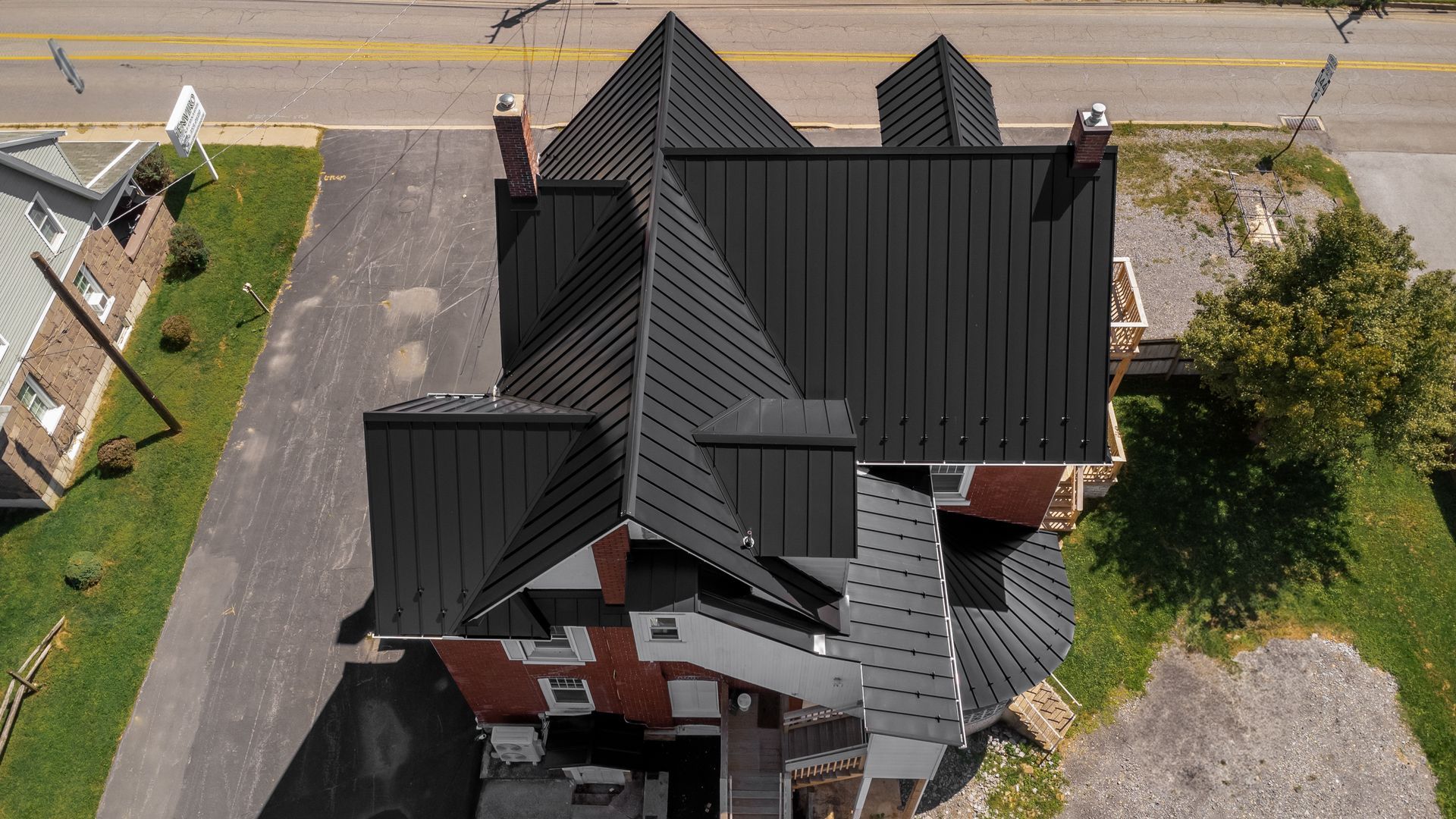 Aerial view of a brick house with a black metal roof, driveway, and surrounding greenery.
