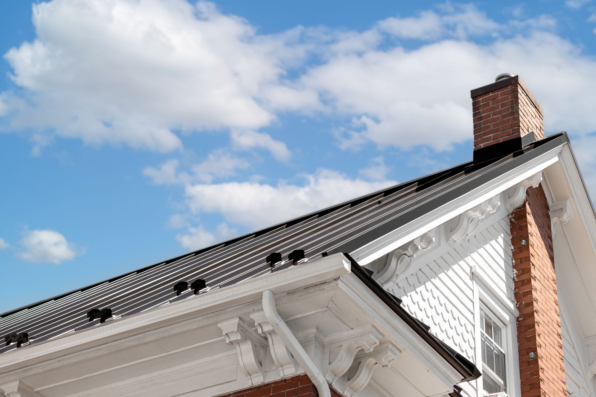 White house roof with dark shingles, brick chimney, and blue sky with clouds.