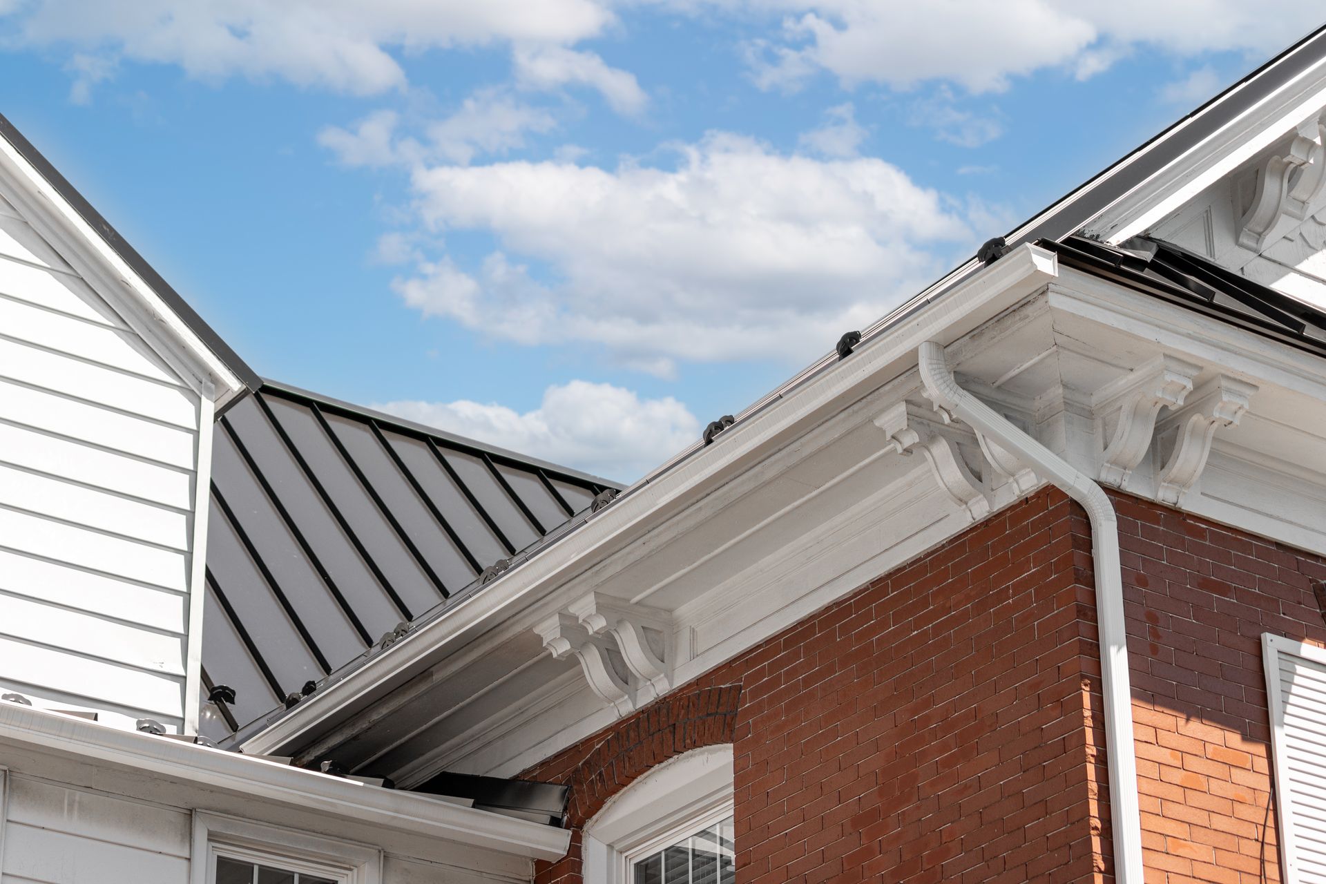 White gutters on a red brick house with a black metal roof against a blue sky.