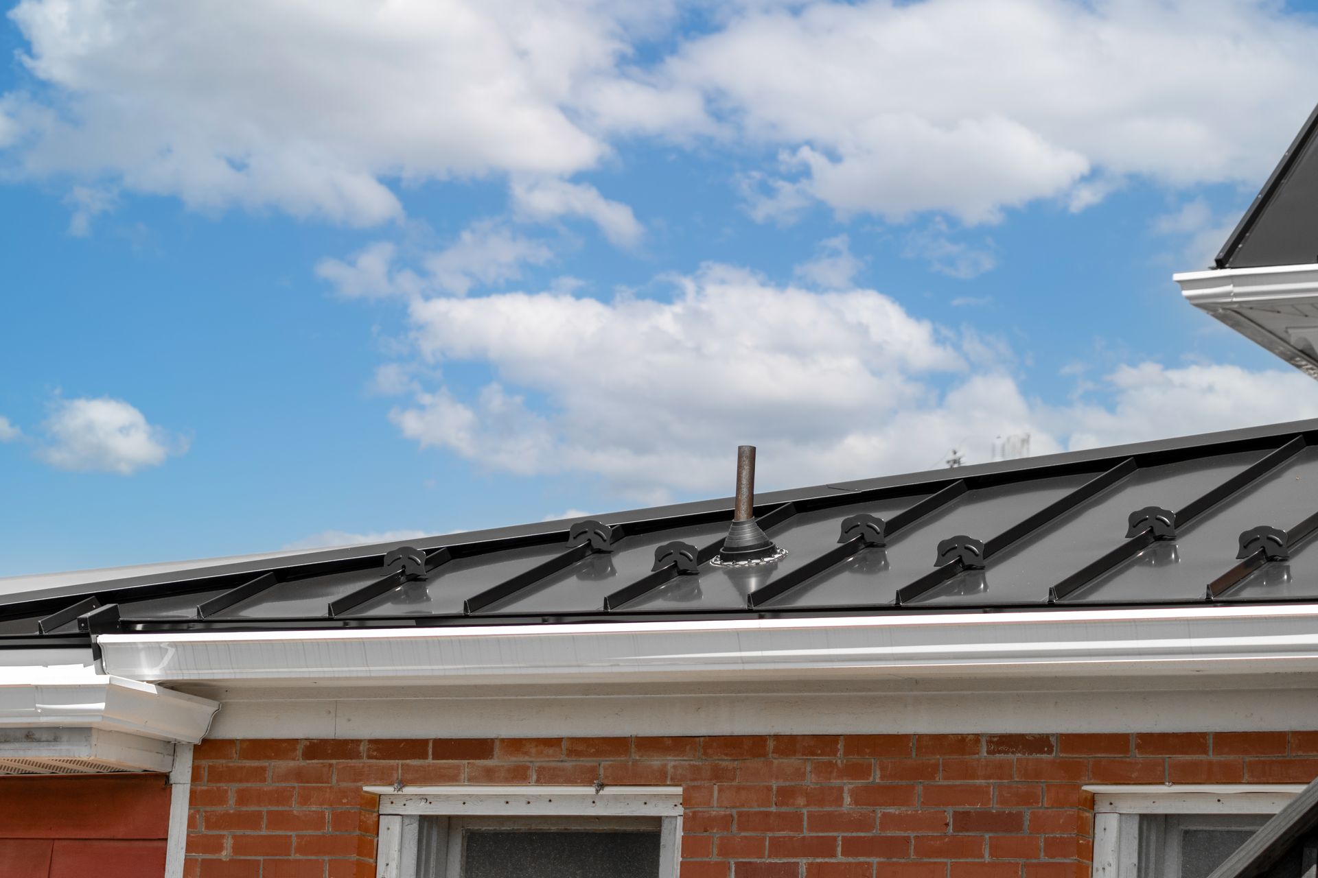 Black metal roof with brick building below, pipe in the center, blue sky with clouds.