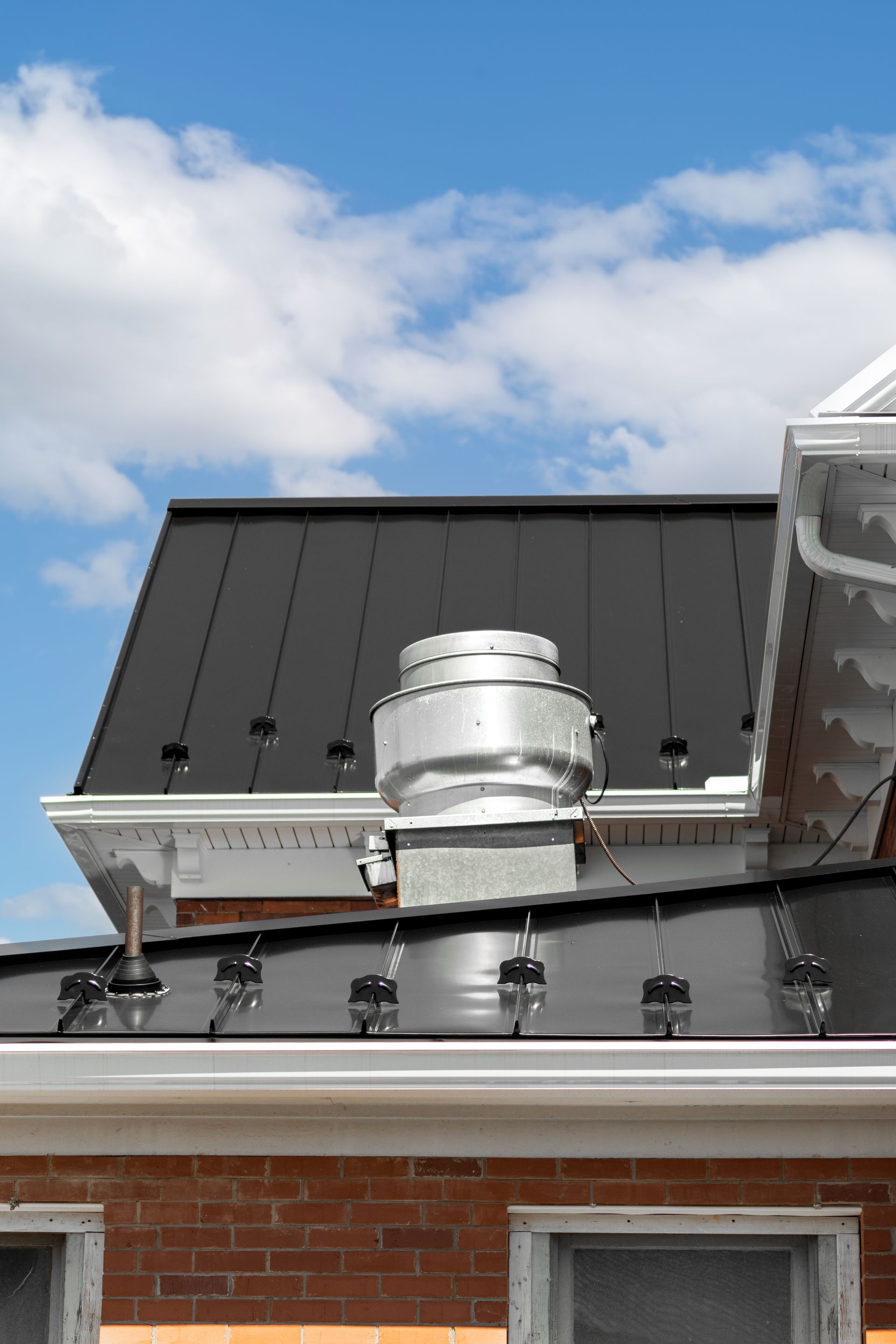 Close-up of a dark roof with a vent and white trim against a blue sky with clouds.