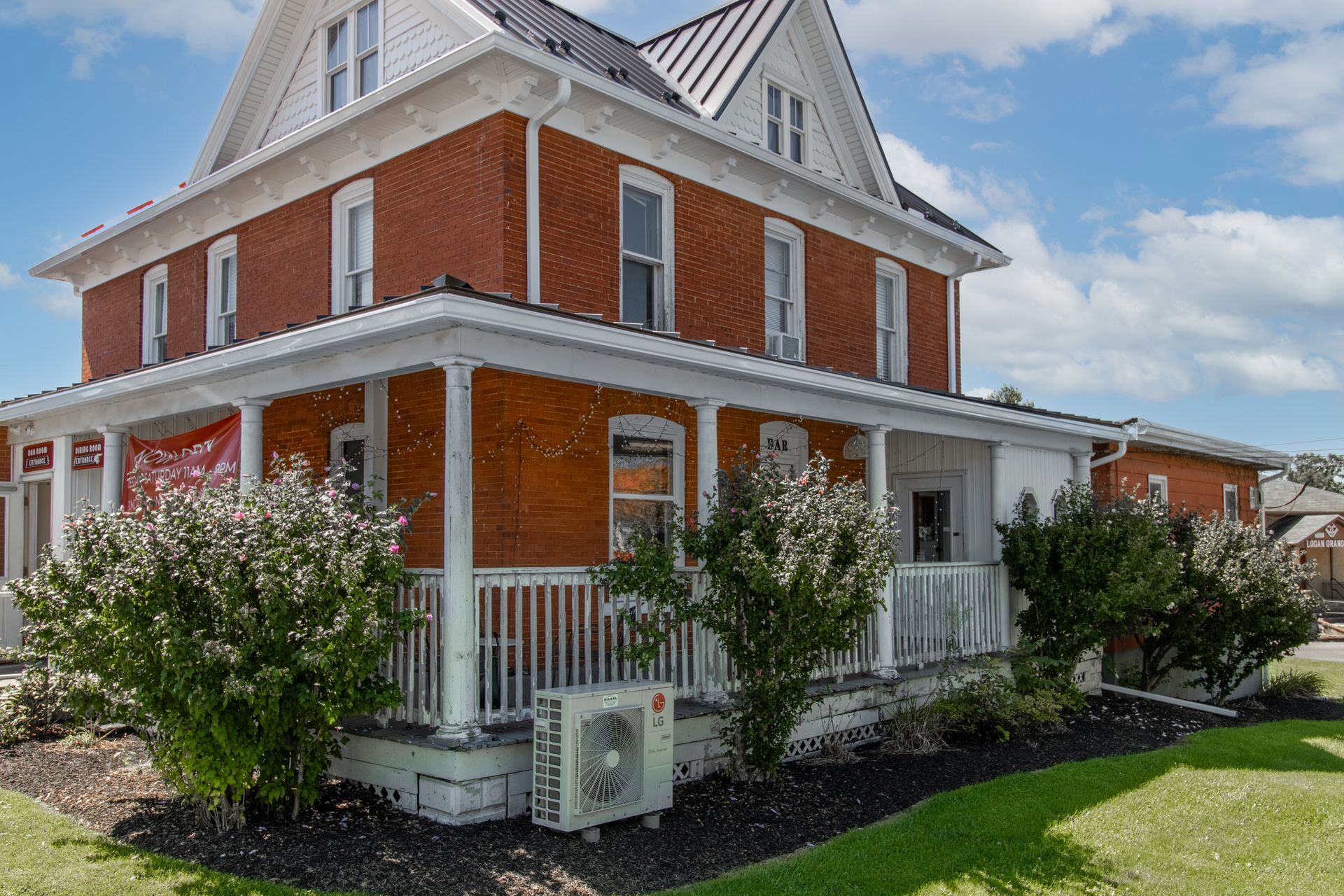 Red brick building with white trim, porch, and bushes under a blue sky.