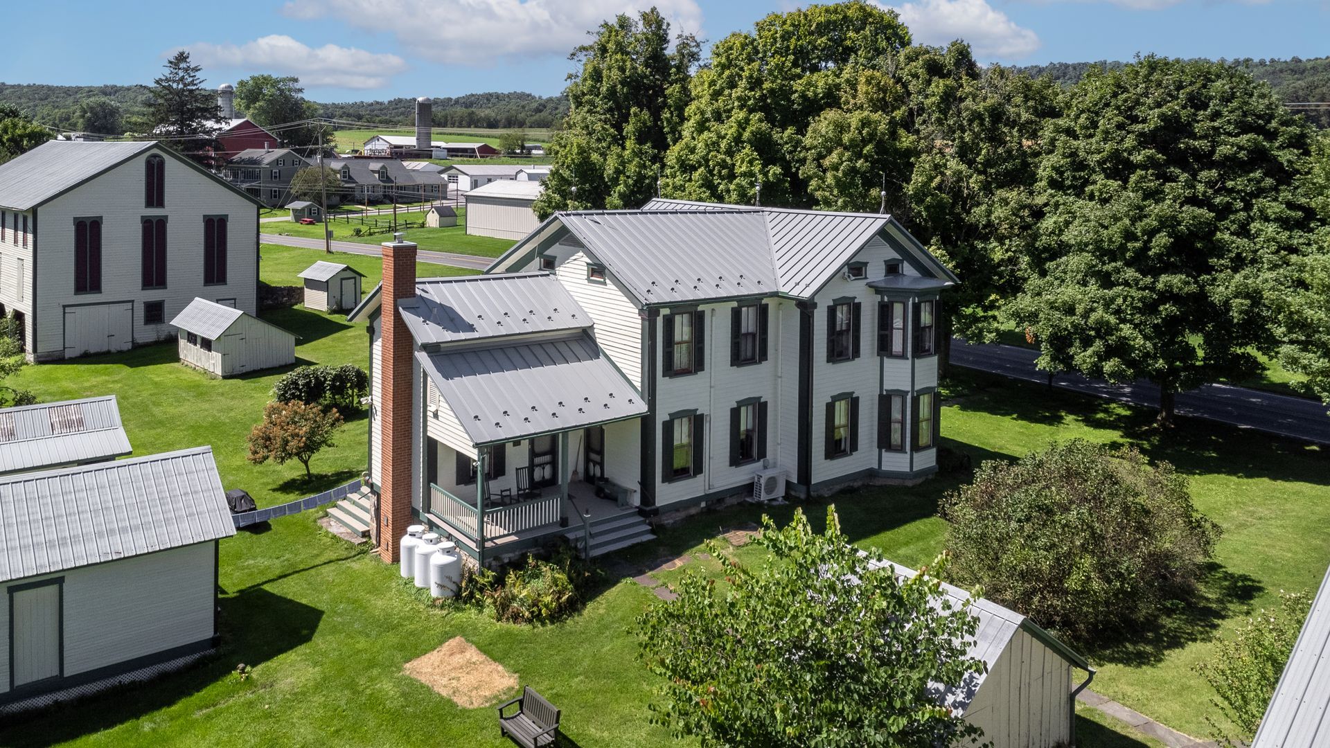White house with a metal roof and porch, surrounded by other small buildings on green grass.