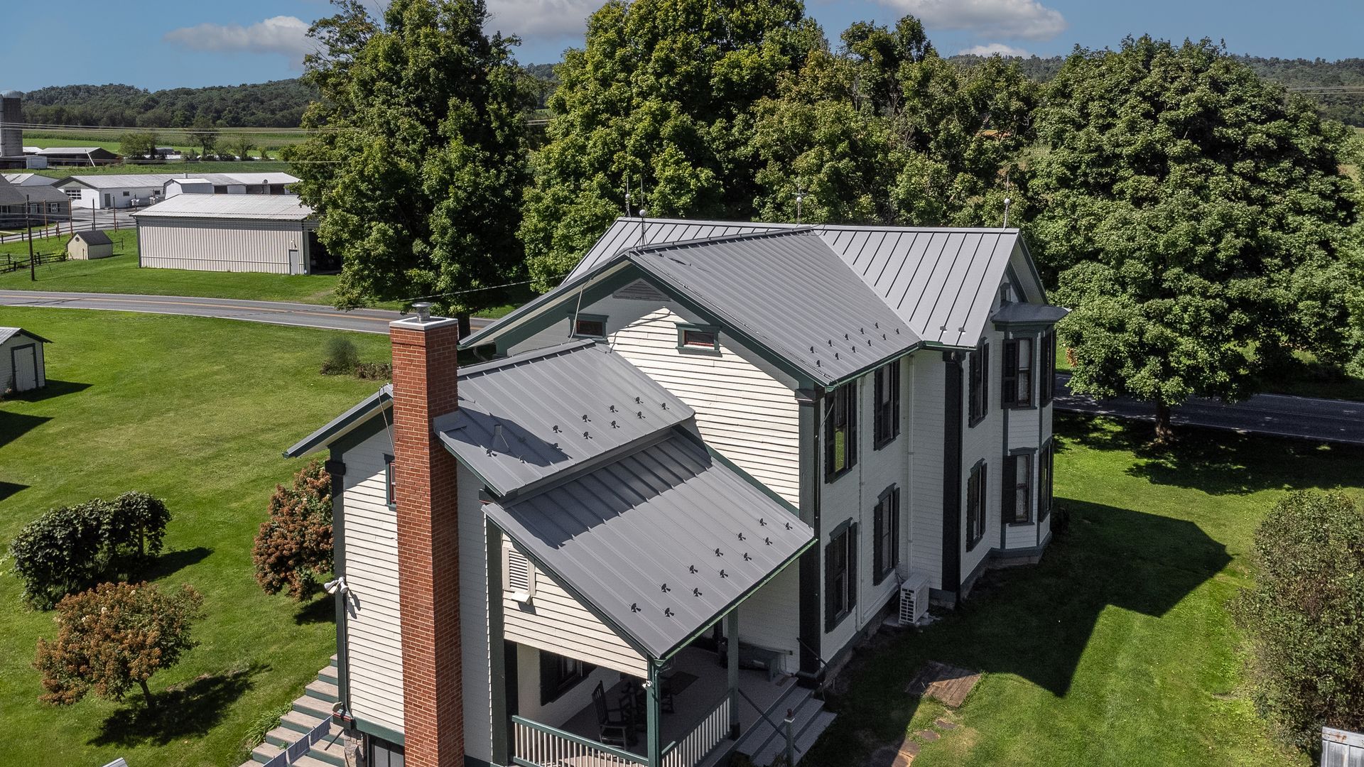 Two-story house with gray metal roof, white siding, black shutters, and chimney on green lawn with trees in the background.