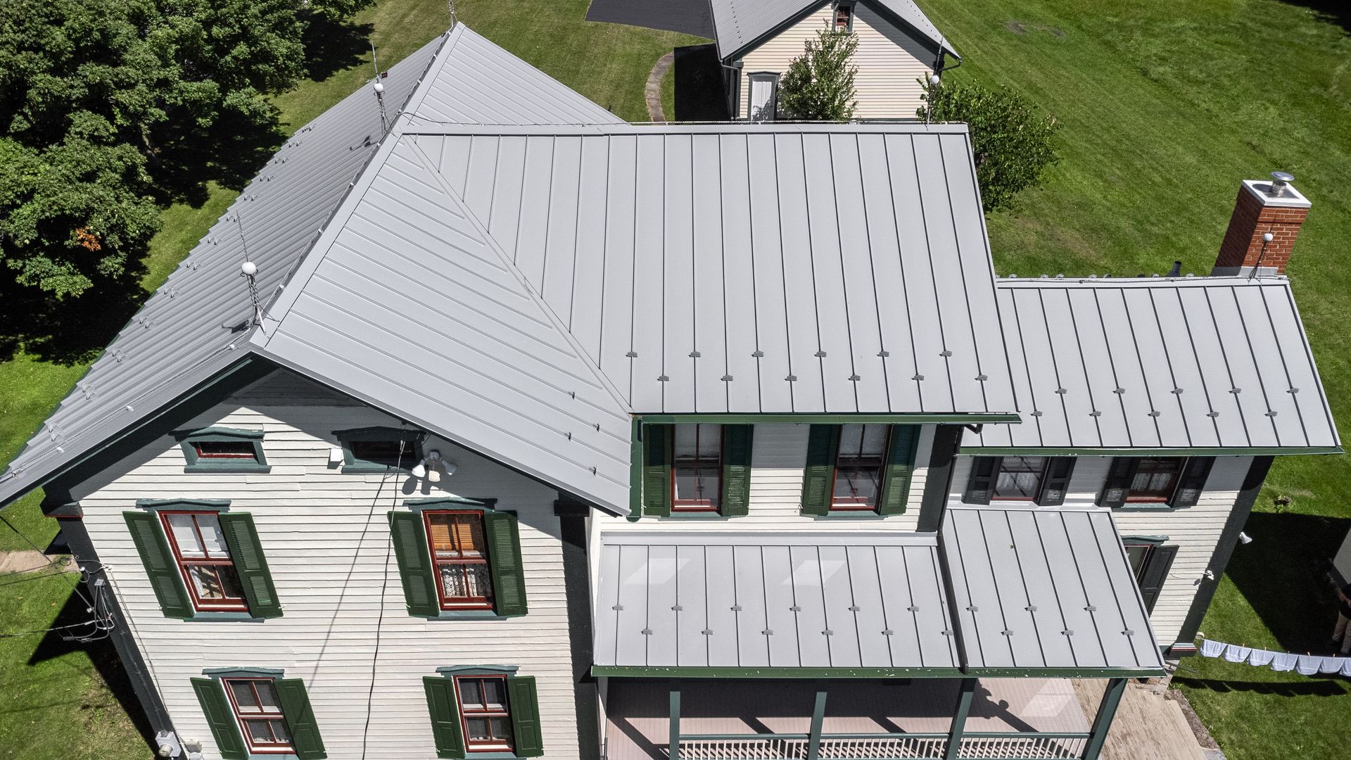Aerial view of a white two-story house with a gray metal roof and green shutters, in a grassy area.
