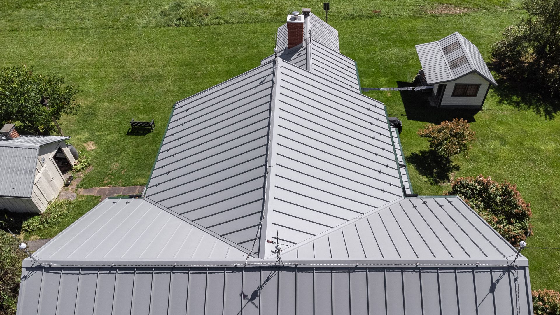 Aerial view of a house with a gray metal roof, chimney, and two outbuildings on green grass.