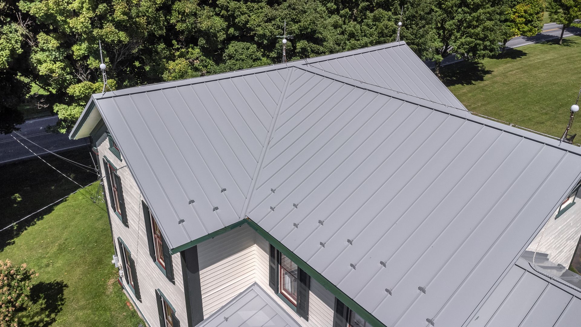 Gray metal roof of a two-story building with black shutters, surrounded by trees and grass.