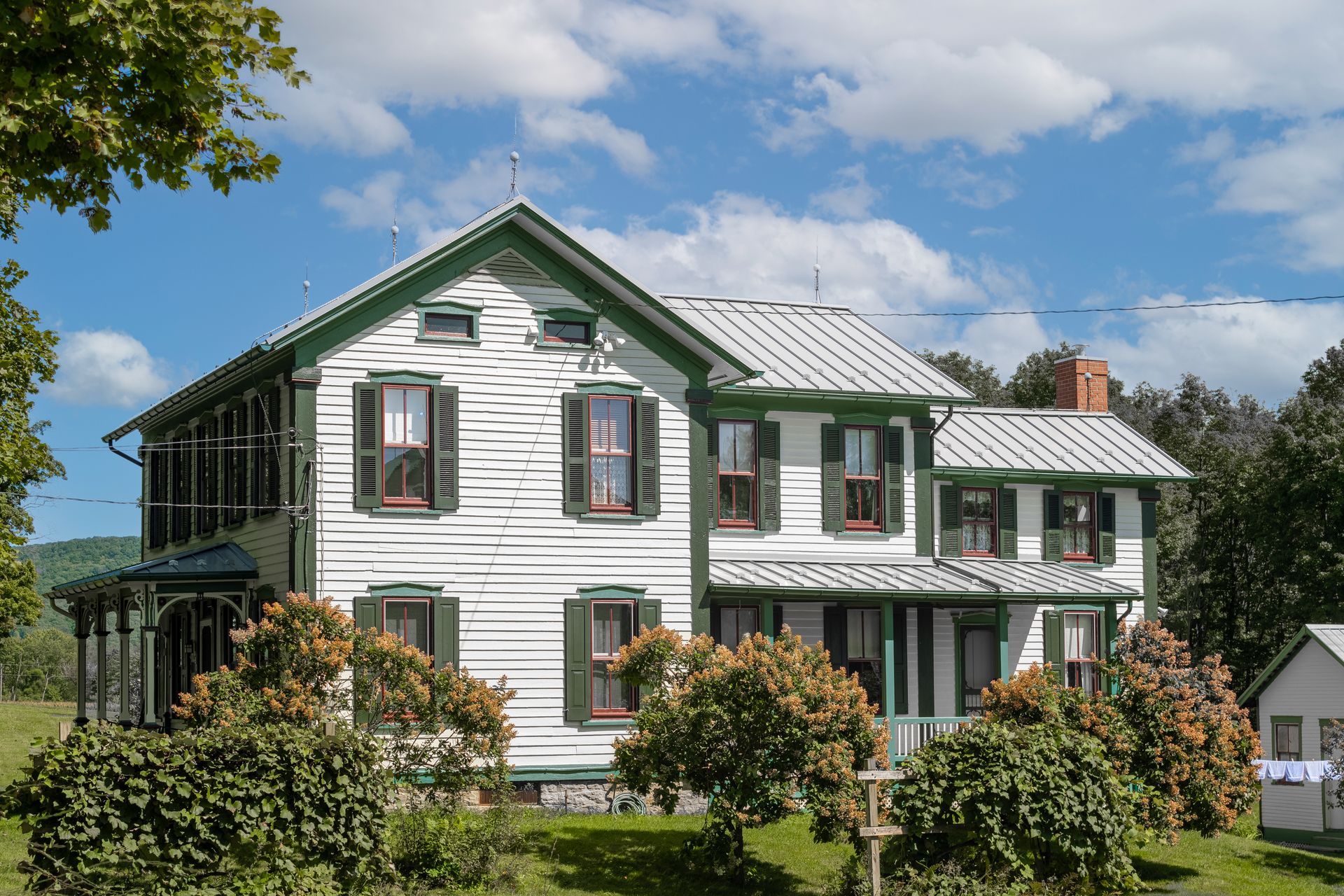White farmhouse with green trim, surrounded by greenery, under a blue sky with clouds.