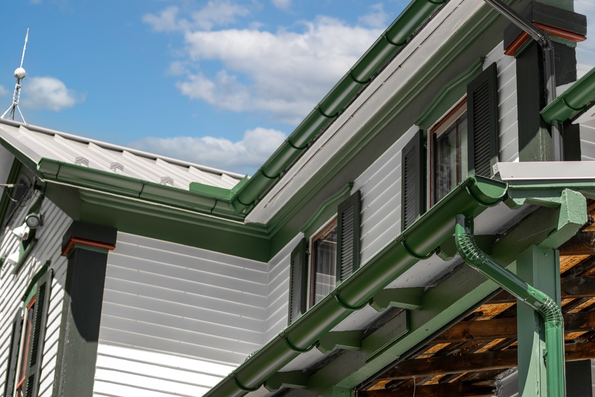 Green gutters on a two-story house with white siding and a metal roof against a blue sky.