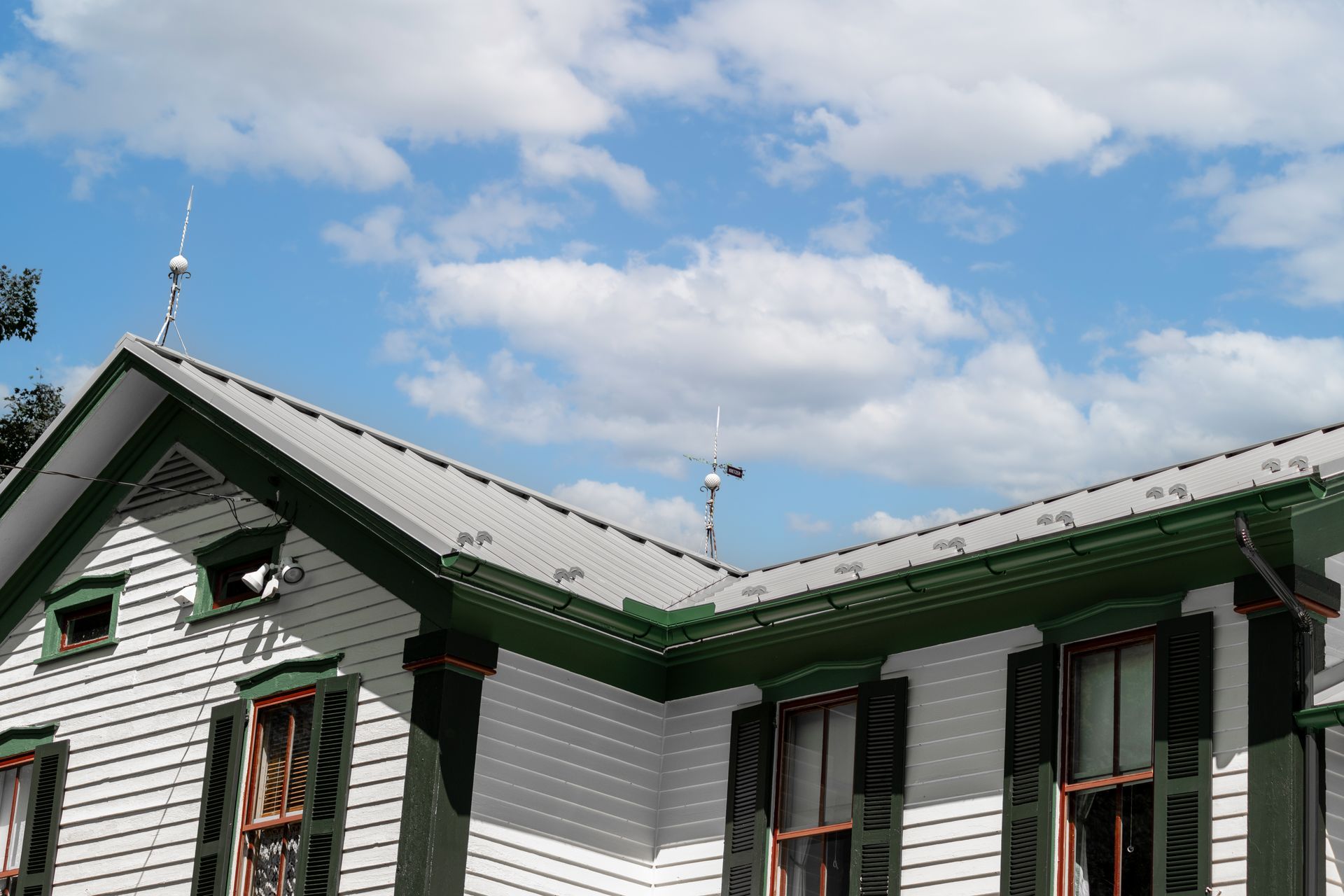 White house with green trim, grey roof, and lightning rods against a blue sky with clouds.