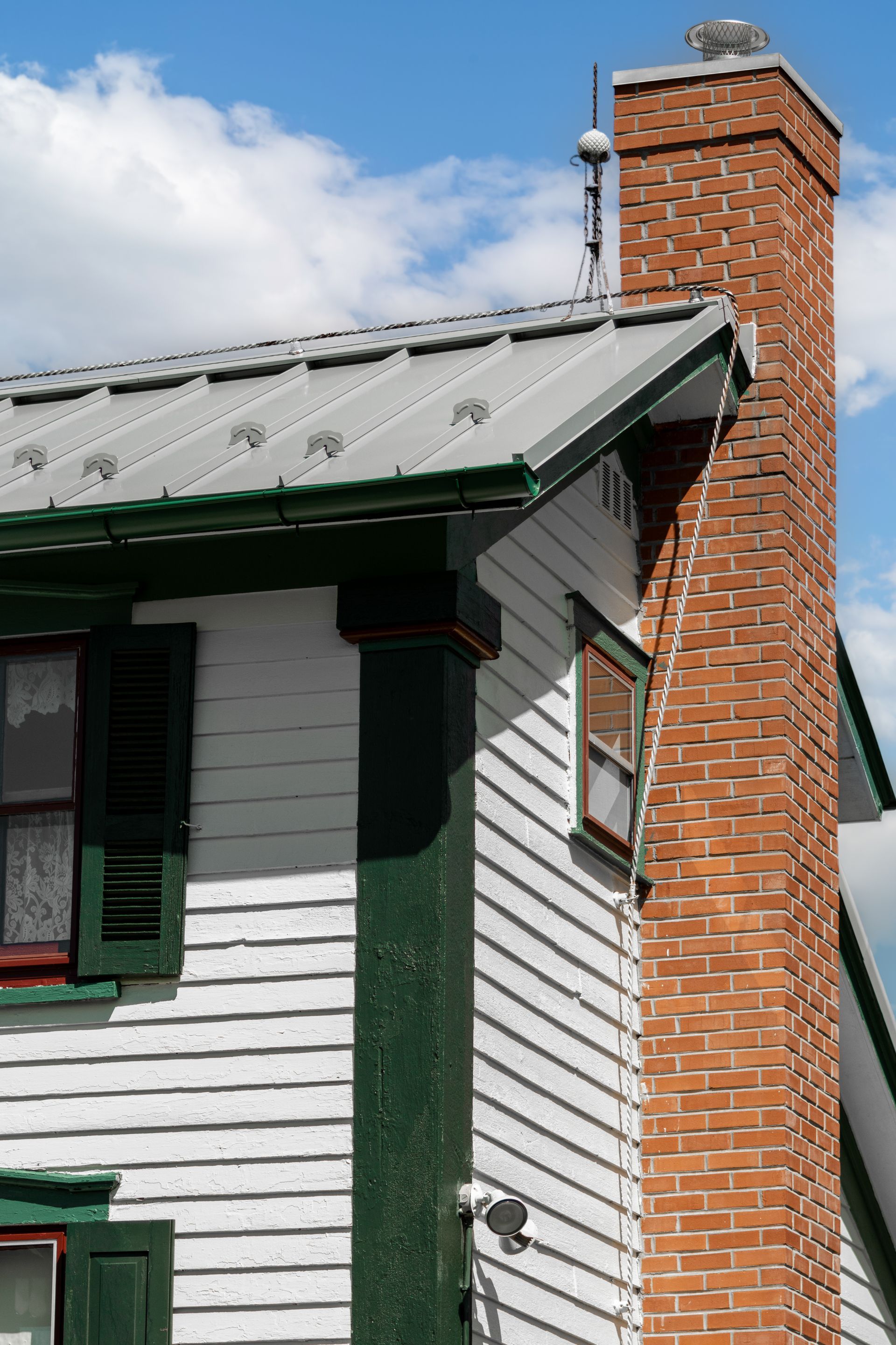White house corner with green trim, metal roof, and red brick chimney against a partly cloudy sky.
