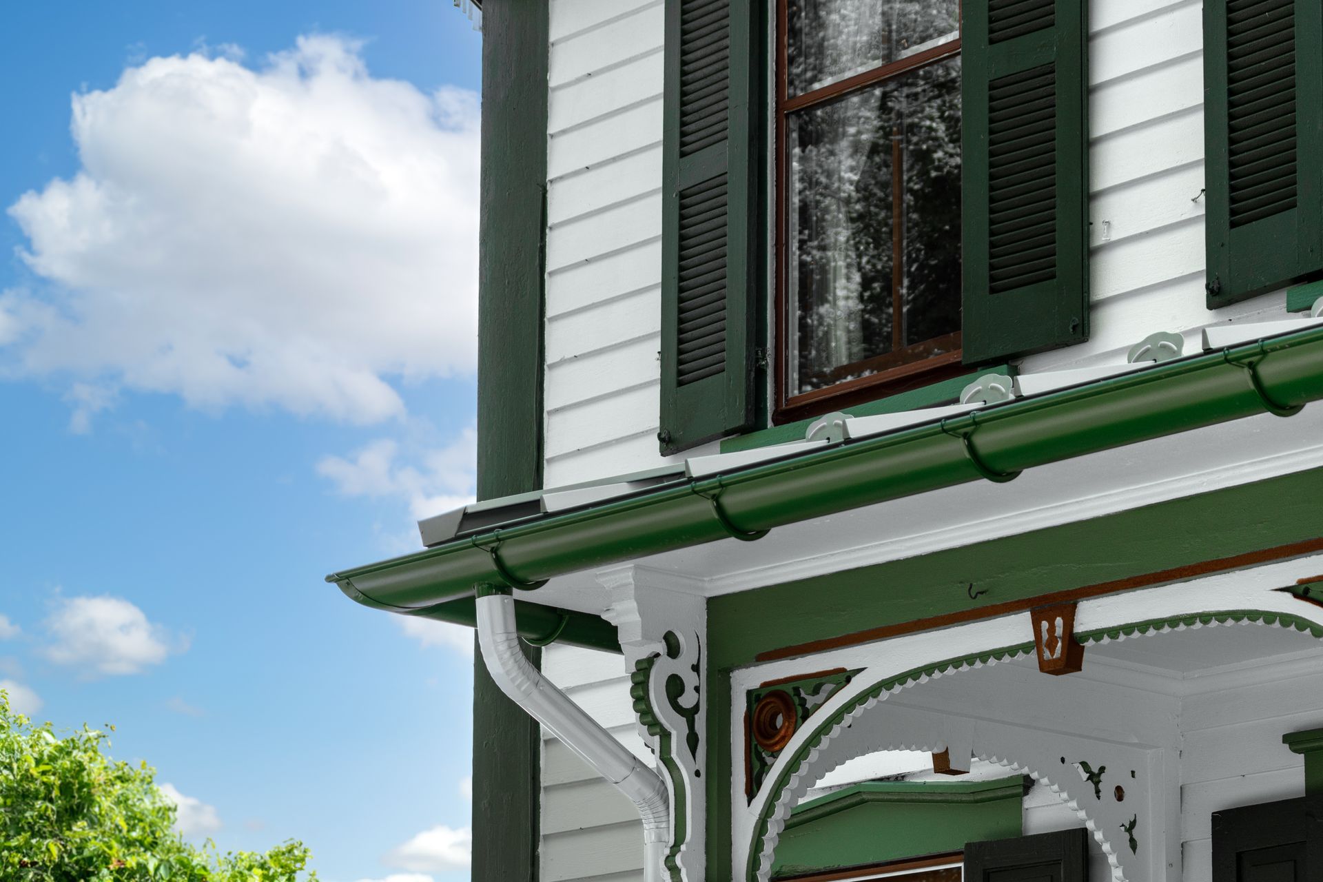 Corner of a white house with green trim, shutters, and gutters against a blue sky with clouds.