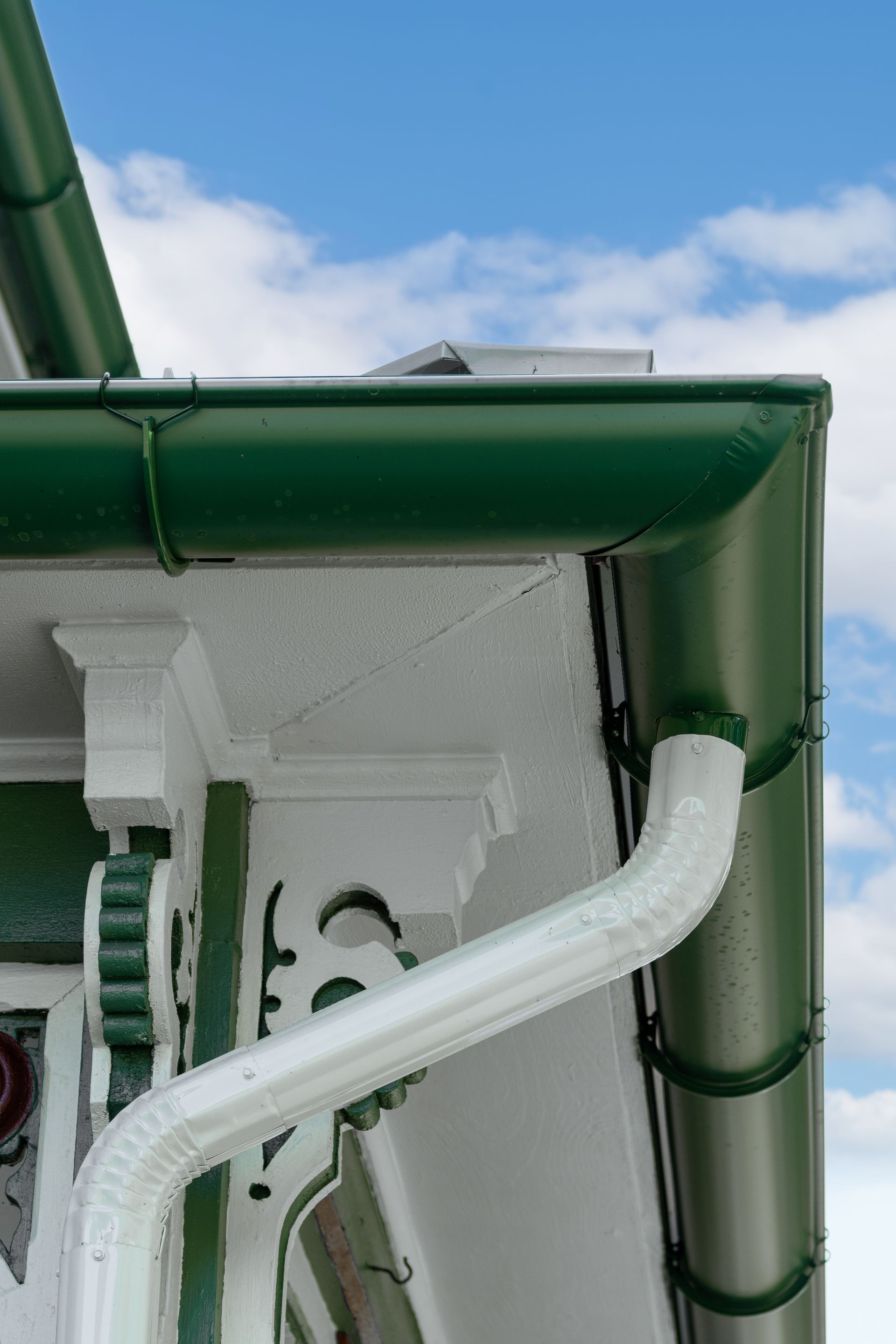Green roof gutter with white downspout, contrasting against a white and green building against a cloudy sky.