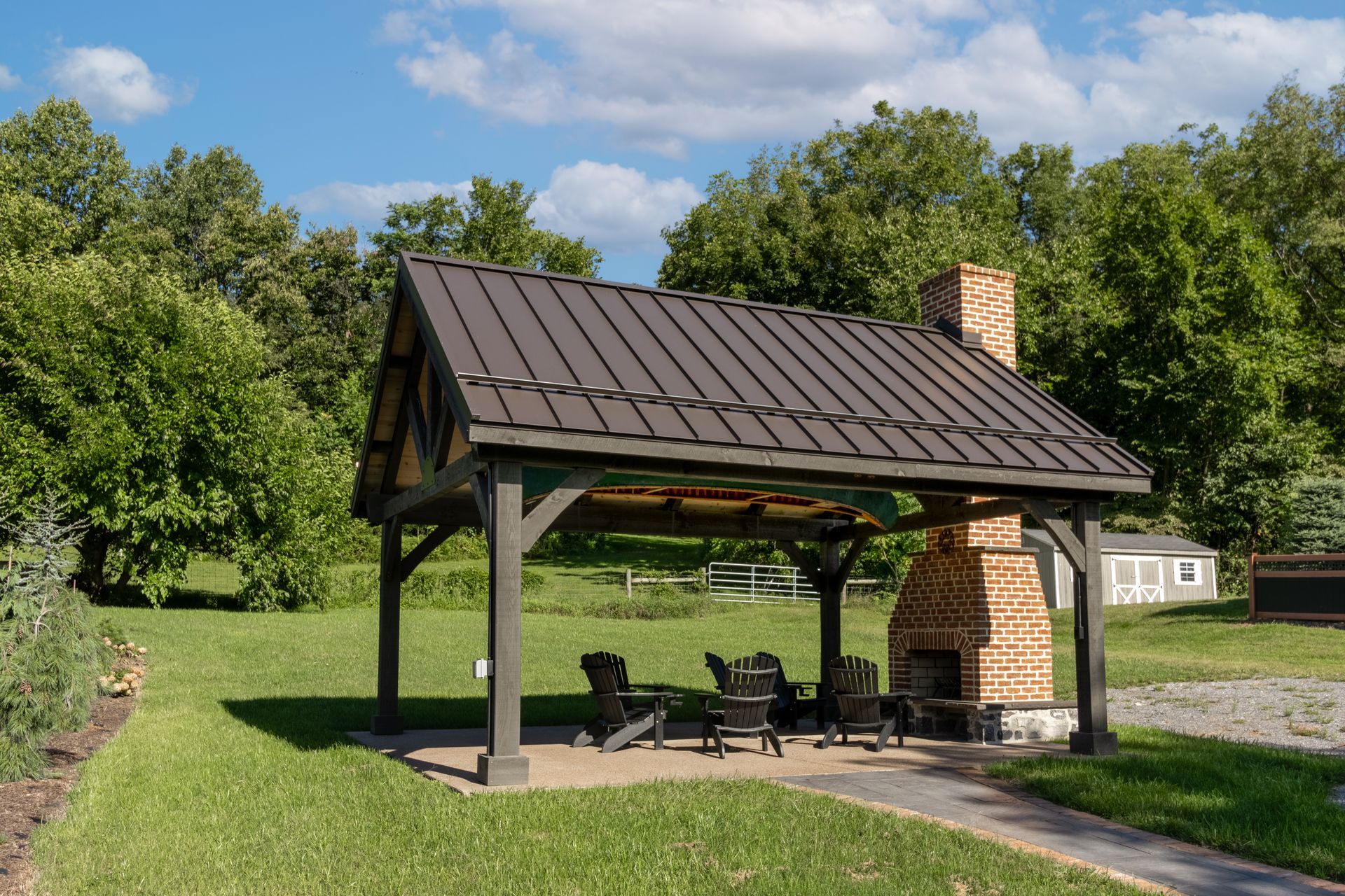 A dark-roofed pavilion with an outdoor fireplace, chairs on a grassy lawn. Trees and a blue sky in the background.
