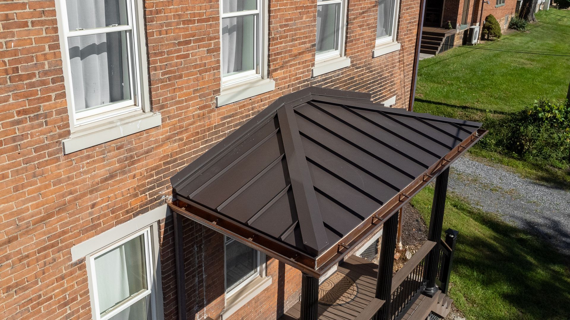 Brown metal roof over a porch on a brick building. White windows and green grass surround.