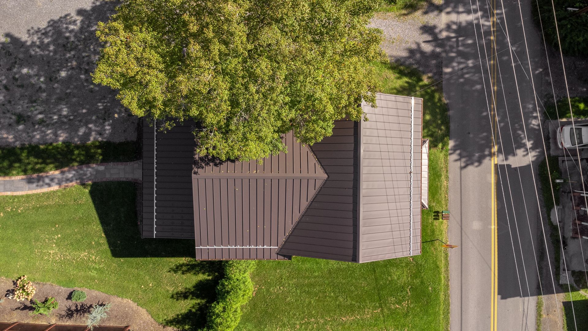 Overhead view of brown-roofed building shaded by a green tree next to a road and grass.