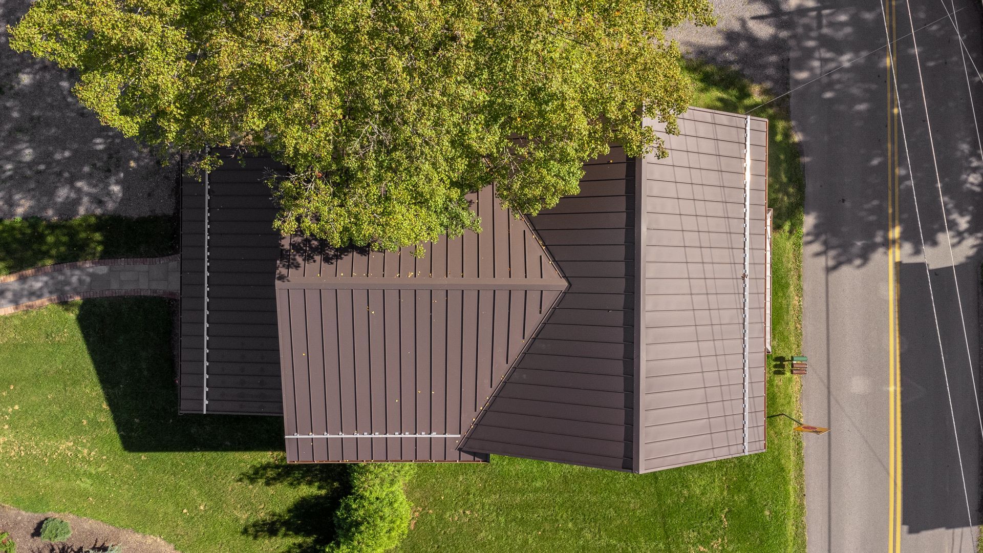 Overhead view of a brown-roofed building with a tree in front and road to the right, green grass.
