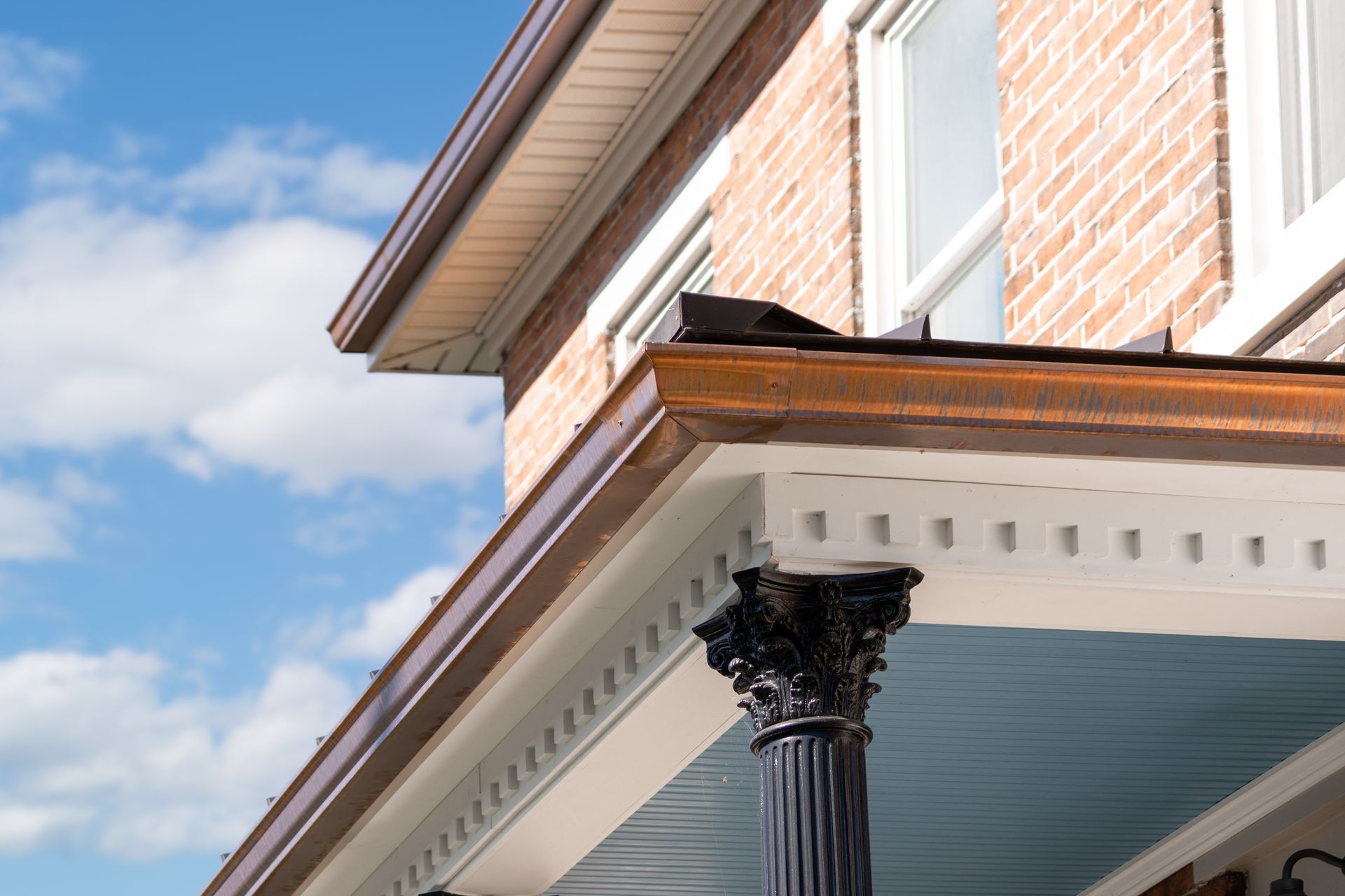 Copper gutters on a brick house, view from below with a decorated porch column against a blue sky.