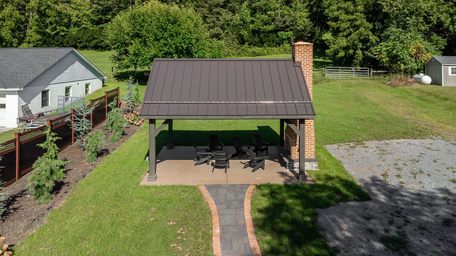 Brown-roofed outdoor pavilion with tables and chairs, chimney, surrounded by green grass and trees.