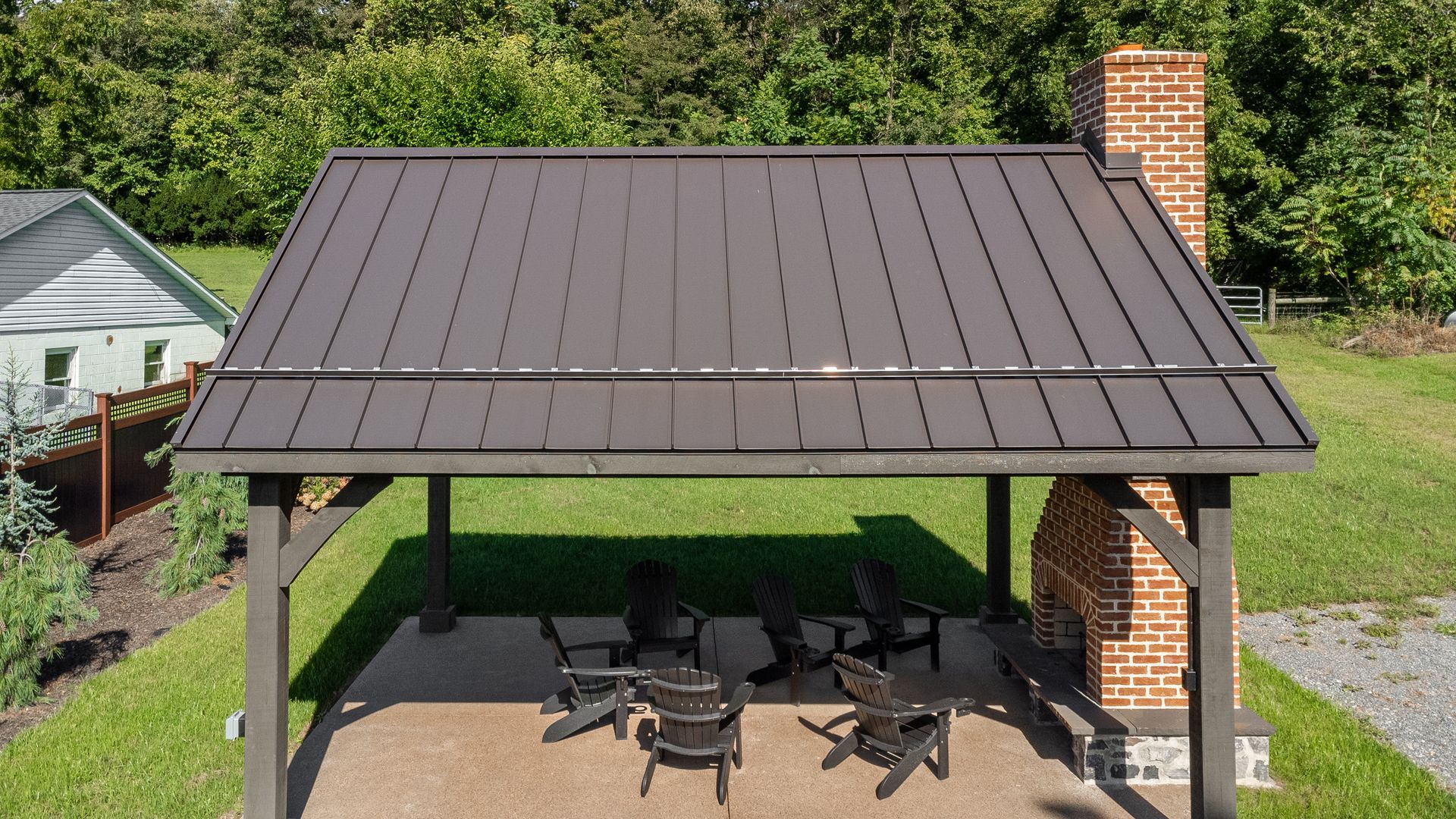 Dark brown metal-roofed pavilion with a fireplace, seating area, and lush green surroundings.