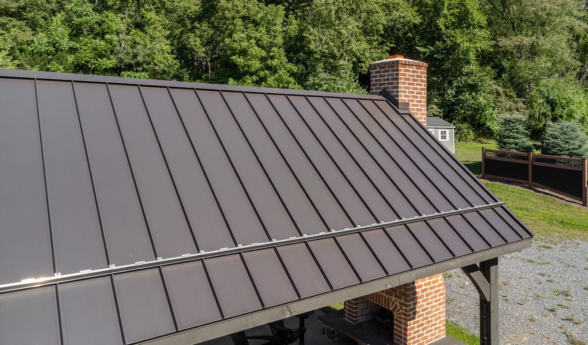 Dark metal roof on a brick building with a chimney. Green foliage in the background.