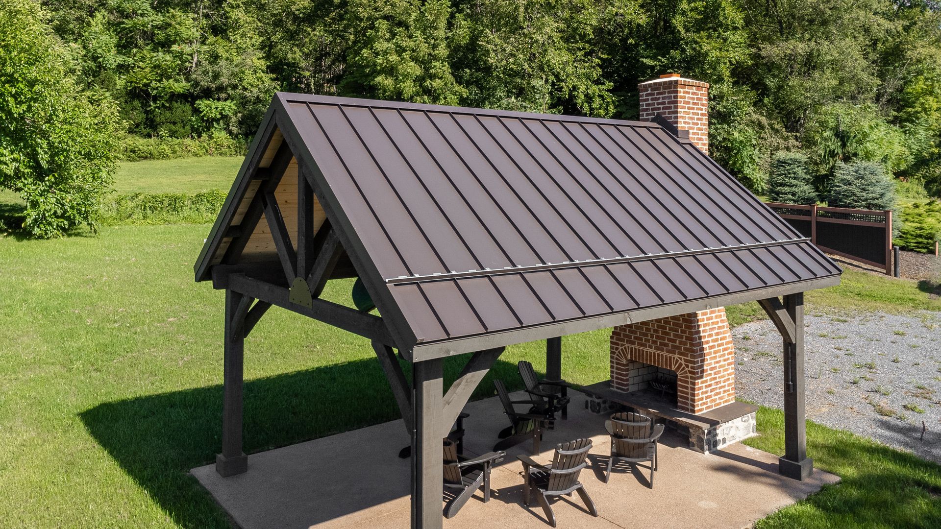A dark brown gazebo with a metal roof, brick fireplace, and outdoor seating in a grassy area.
