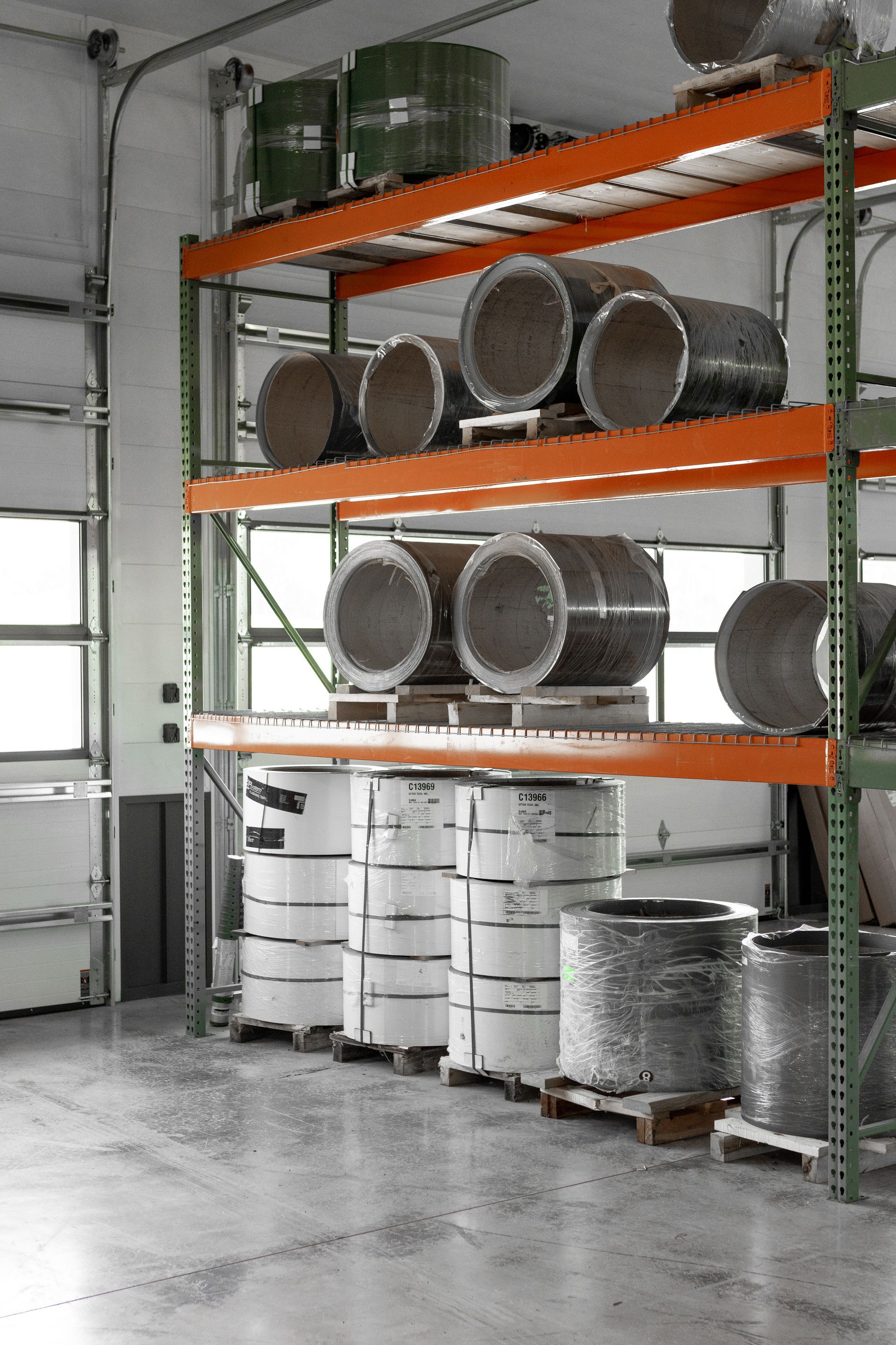 Shelves stocked with large rolls of metal in a warehouse setting, near a loading door.