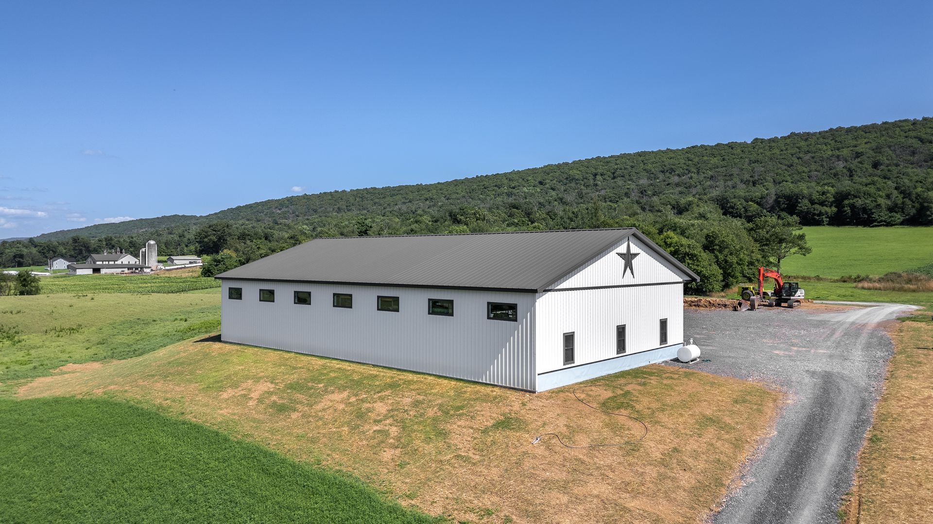 White barn with a star, on a hill. Green field, mountains, and a dirt road are visible.