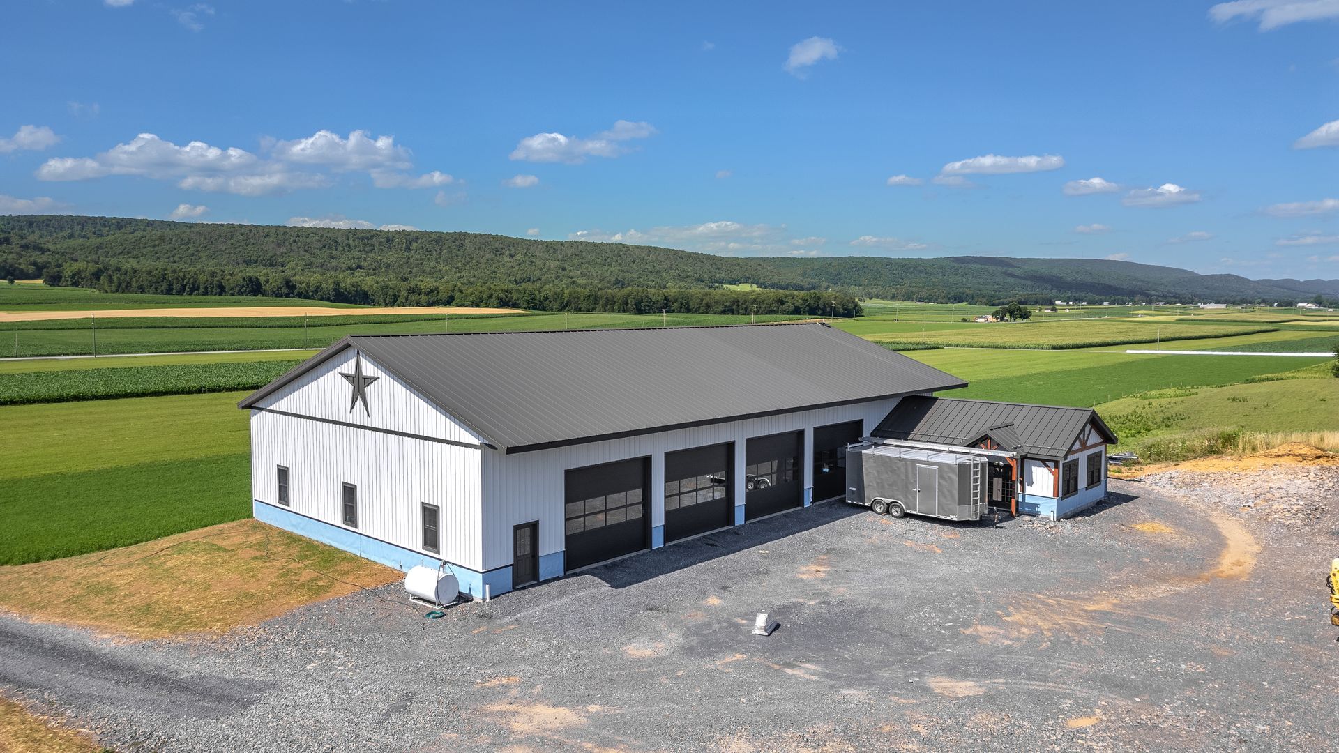 White building with black roof and garage doors, set in a rural landscape with fields and hills.