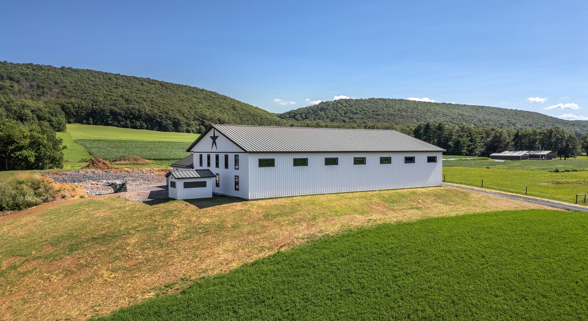 White barn with a dark roof stands in a field with hills in the background under a blue sky.
