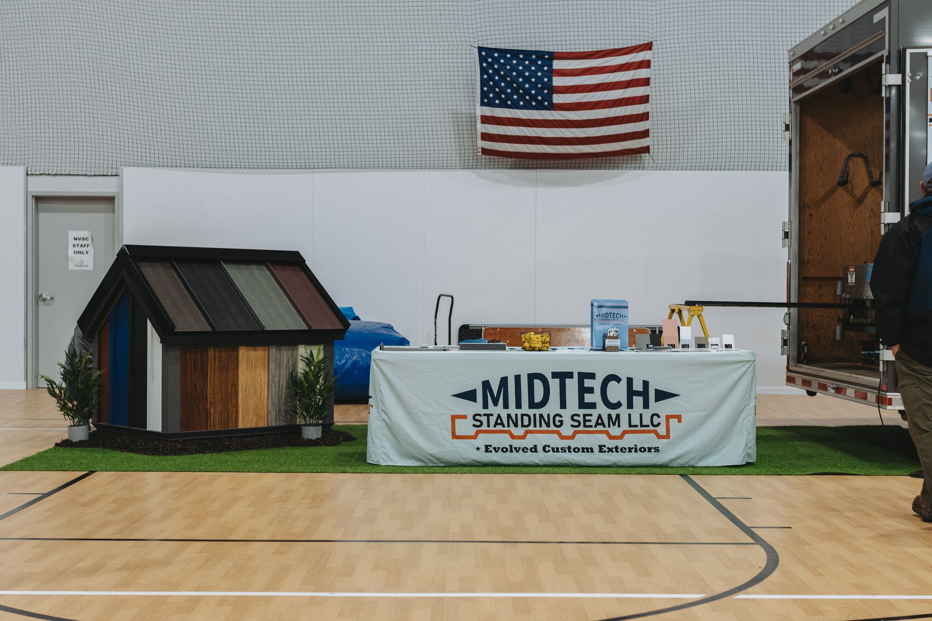 Booth with Midtech Standing Seam LLC sign, miniature roof display, American flag, and trailer in a gym.