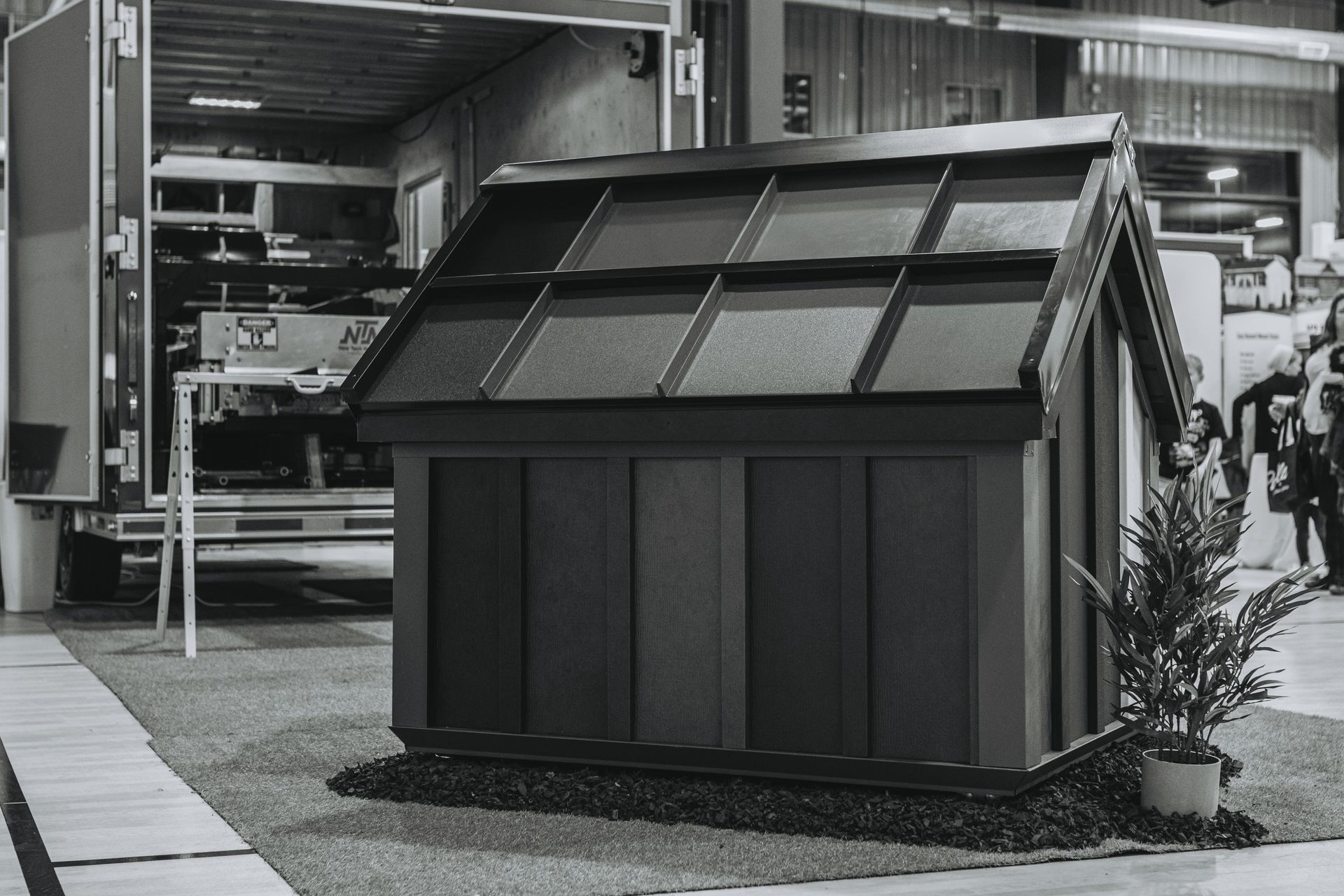 Black and white photo of a small shed with a pitched roof, possibly at a trade show.