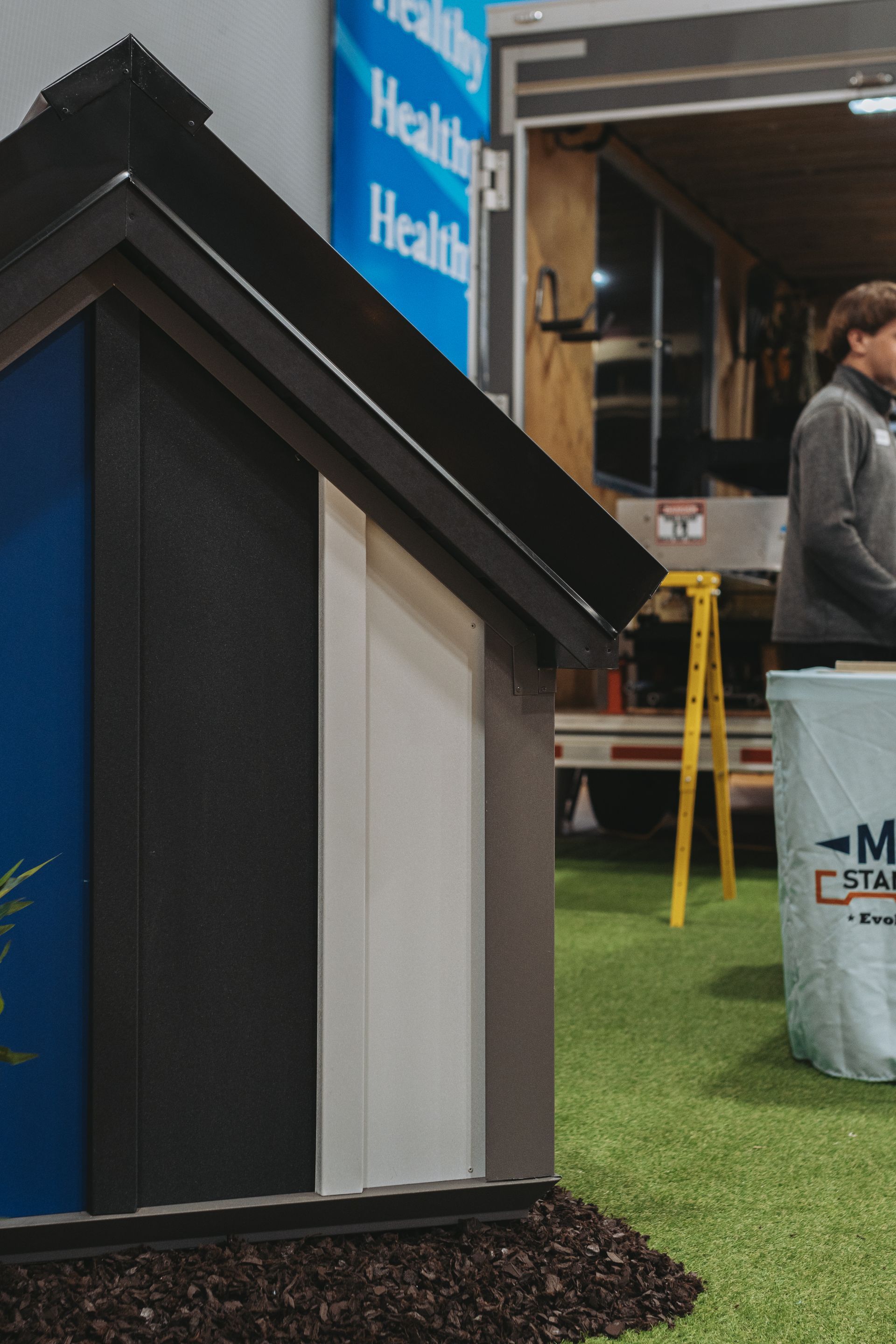 A miniature house model with blue, black, white, and grey siding stands on artificial turf.