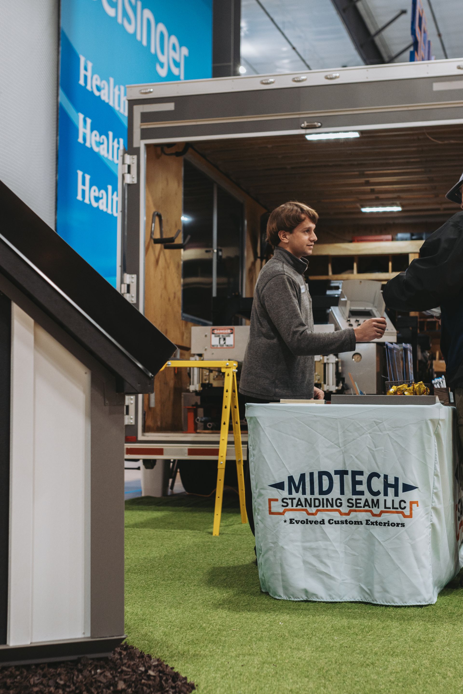 Woman at a trade show booth, Midtech Storage, offering a sample. A model shed is in the foreground.