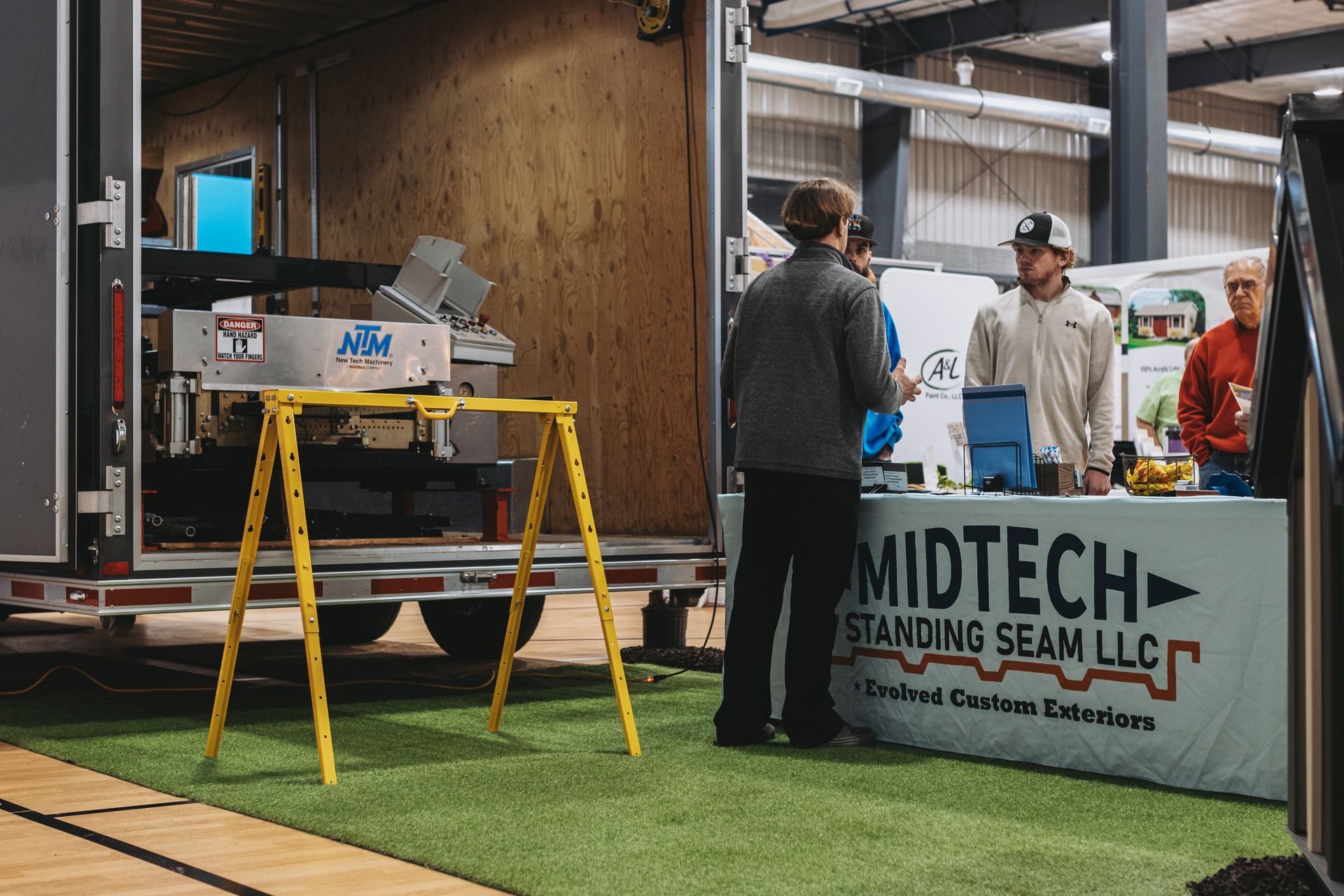 People at a trade show booth for Midtech Standing Seam LLC, with a machine and artificial turf.