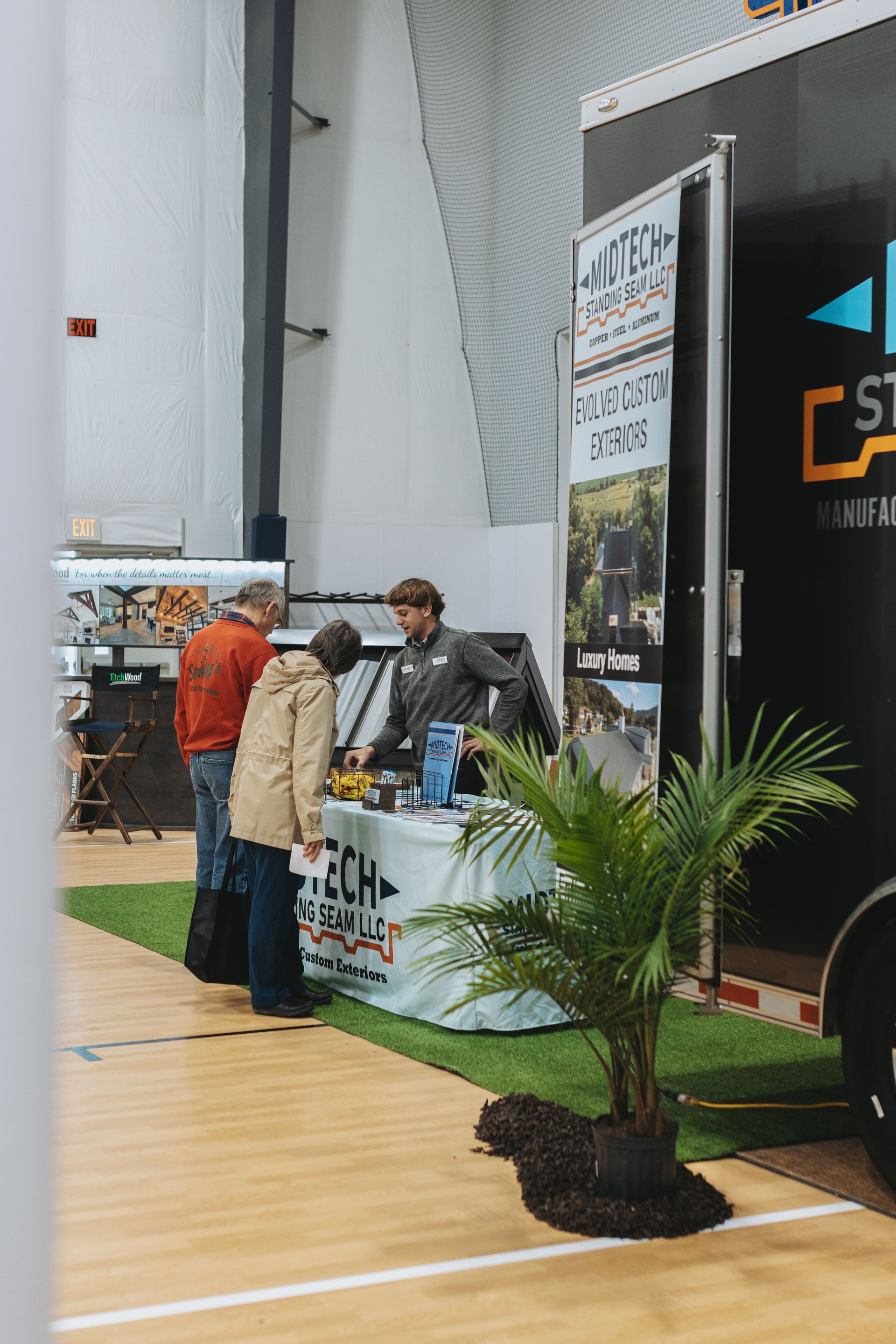 People at a trade show booth with a table, a plant, and a trailer.