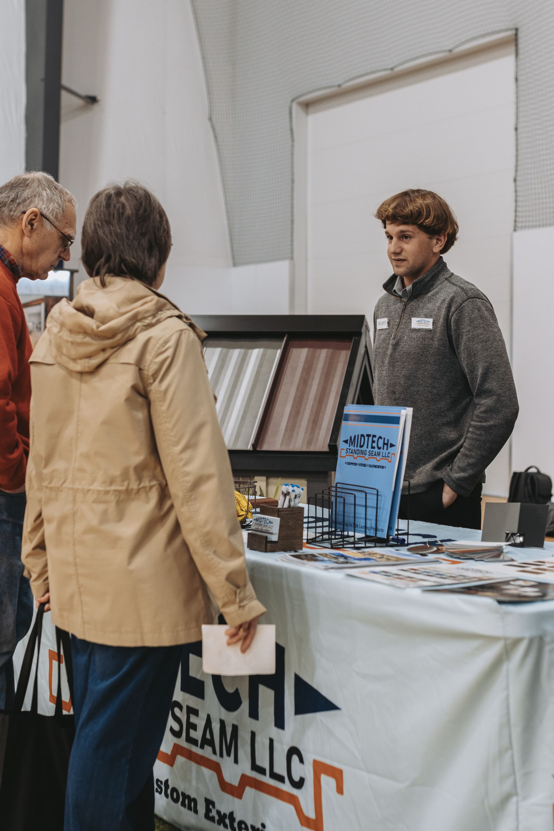 A man at a booth with a woman, looking at roofing samples. Sign reads 
