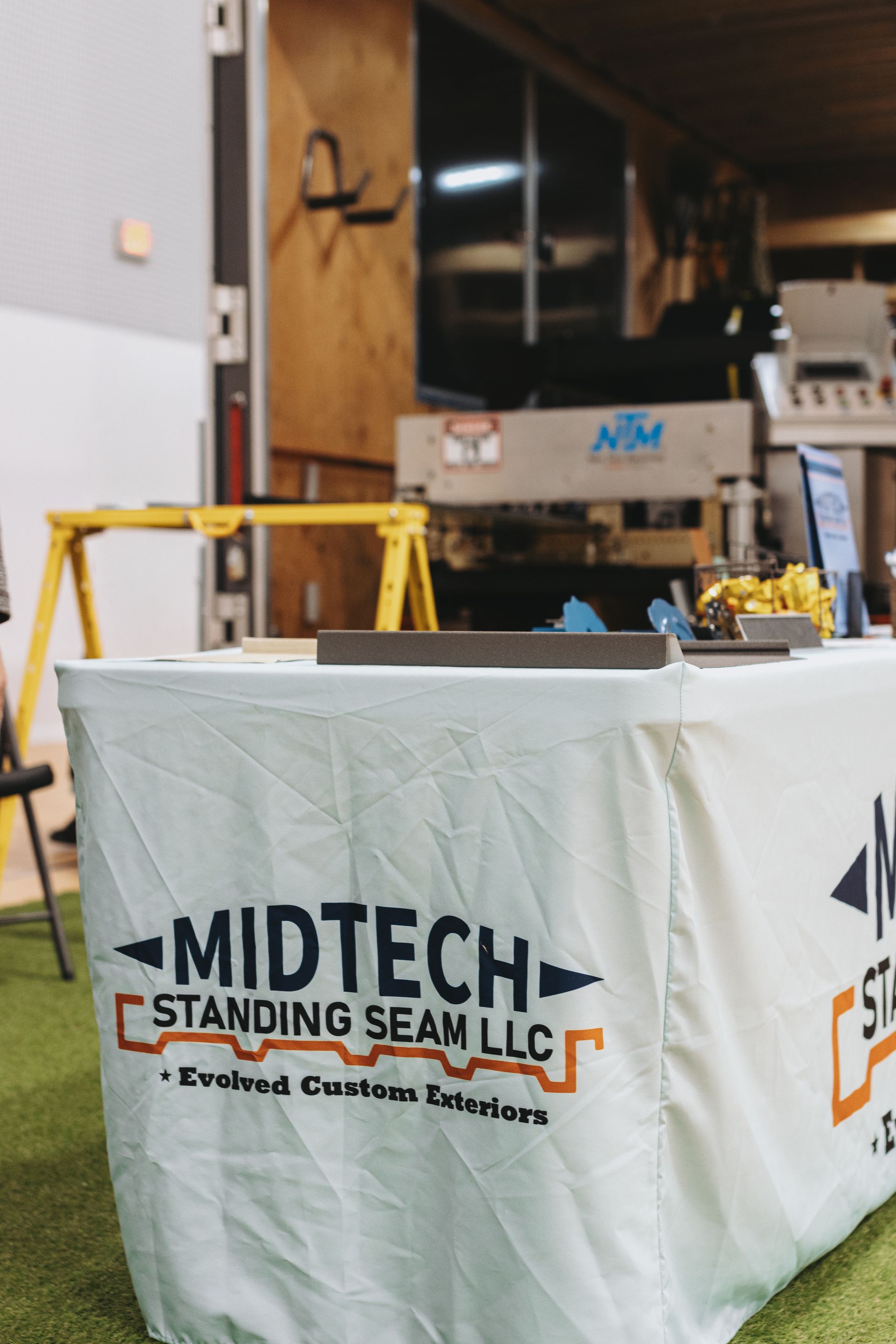 Midtech Standing Seam LLC table with logo in front of machinery at an event.