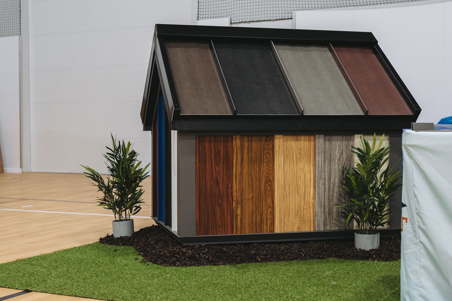Small house model with various wood-tone roof and wall panels on a green lawn.