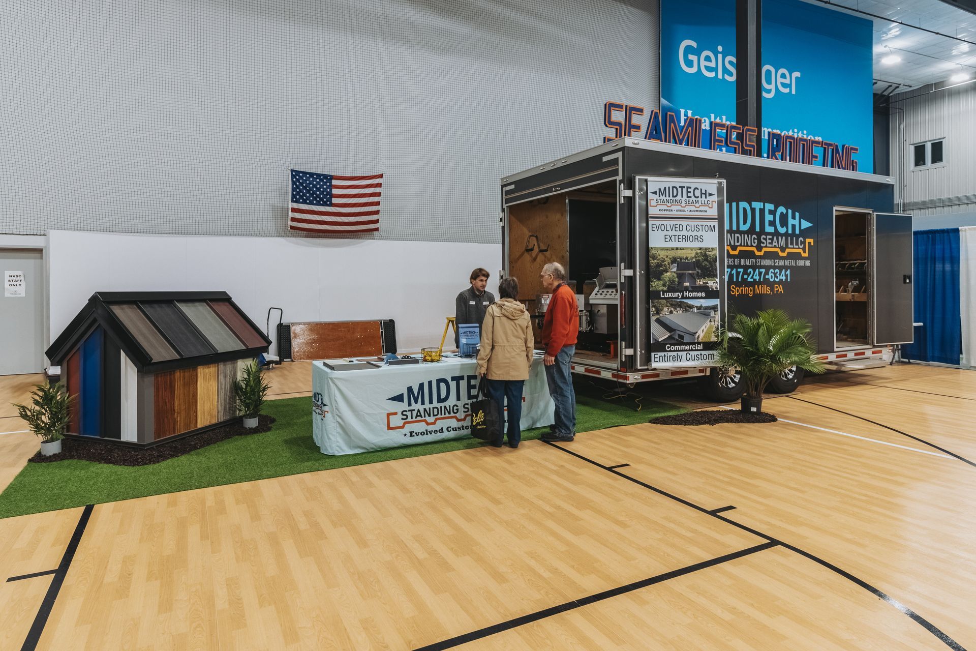 Trade show booth with people, trailer, roofing samples, American flag, and artificial grass.