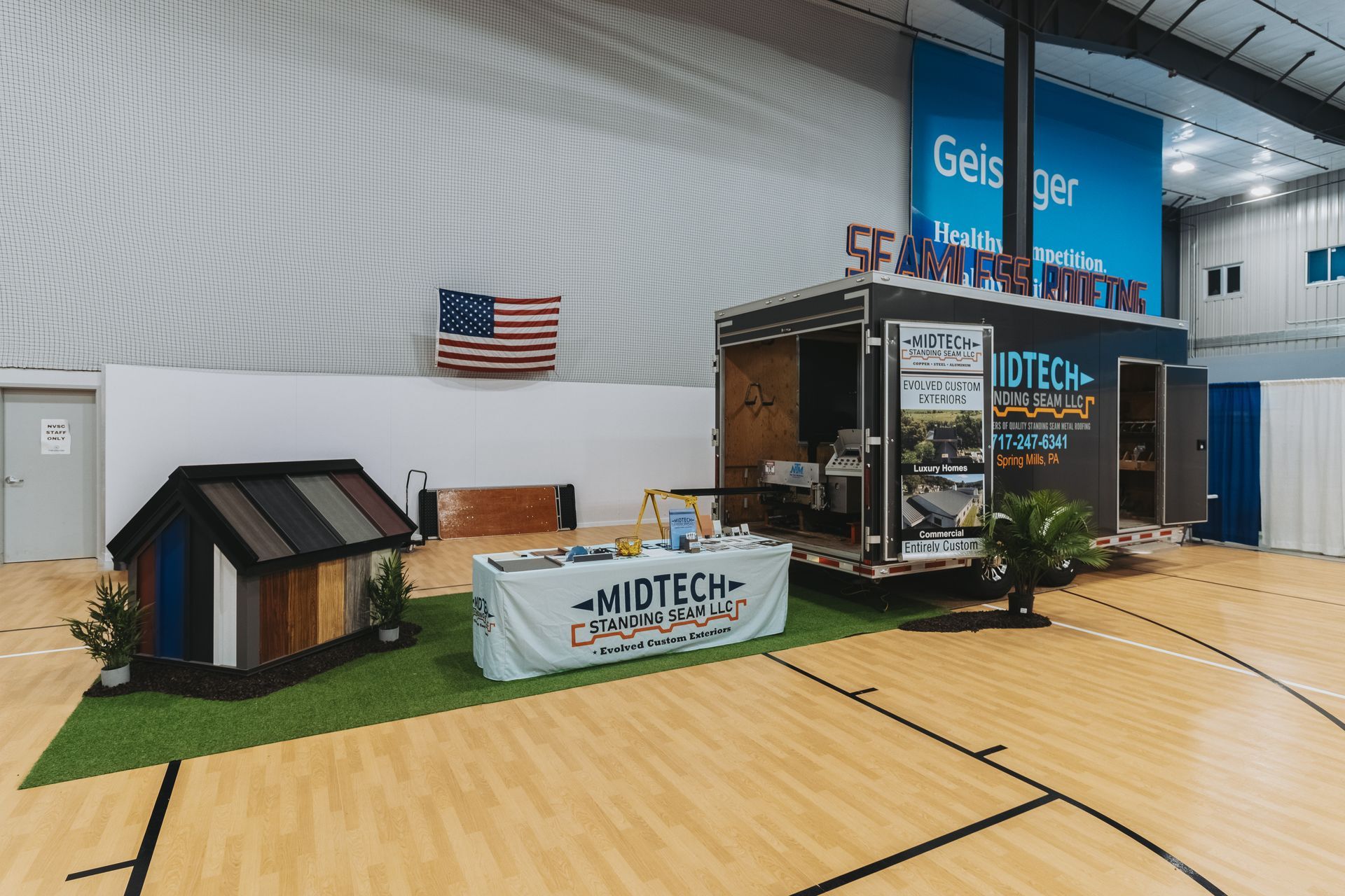 Trade show booth with a small house model, US flag, and signage on a basketball court.