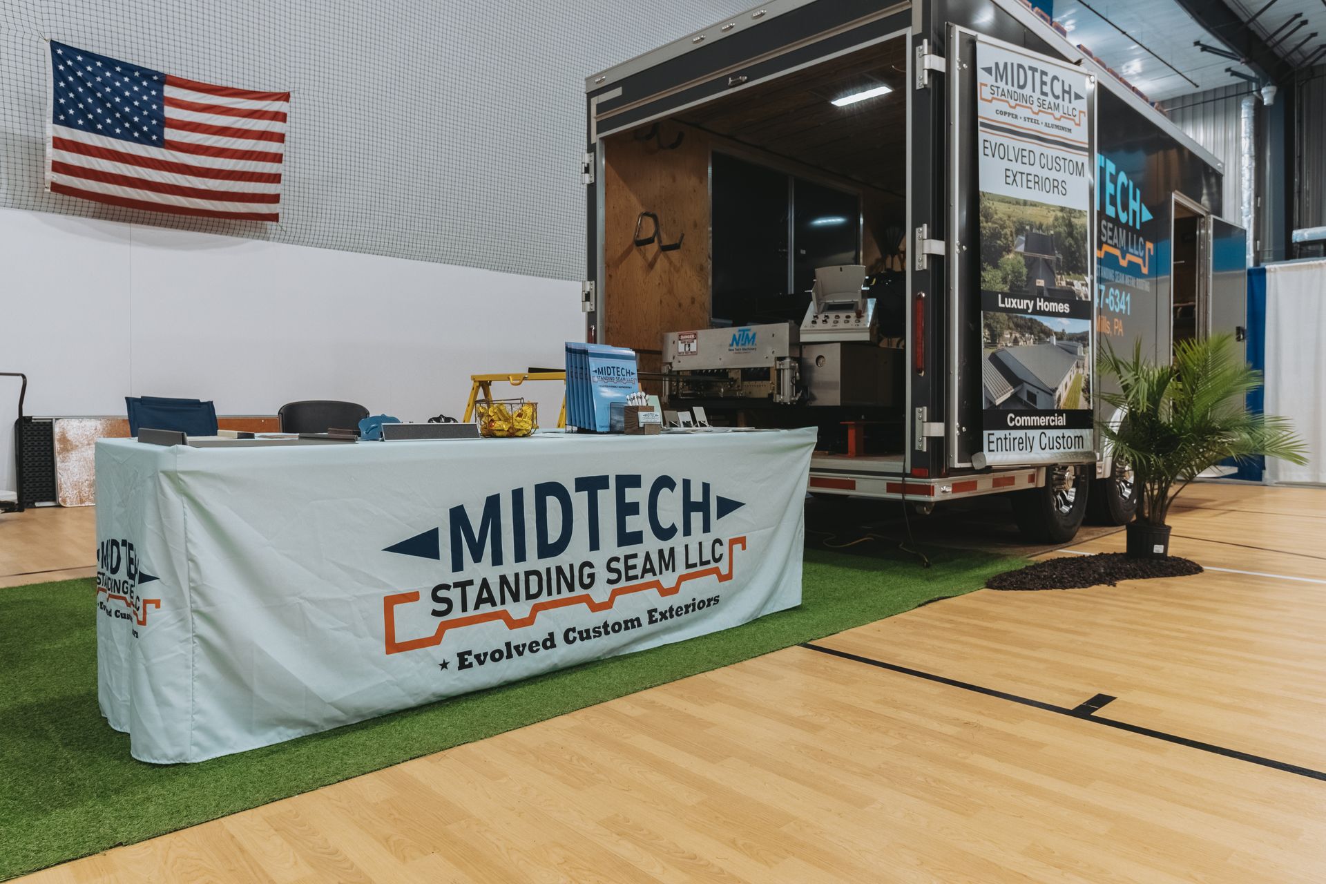 Midtech Standing Seam LLC trade show booth with trailer, table, US flag, and turf on a wooden floor.