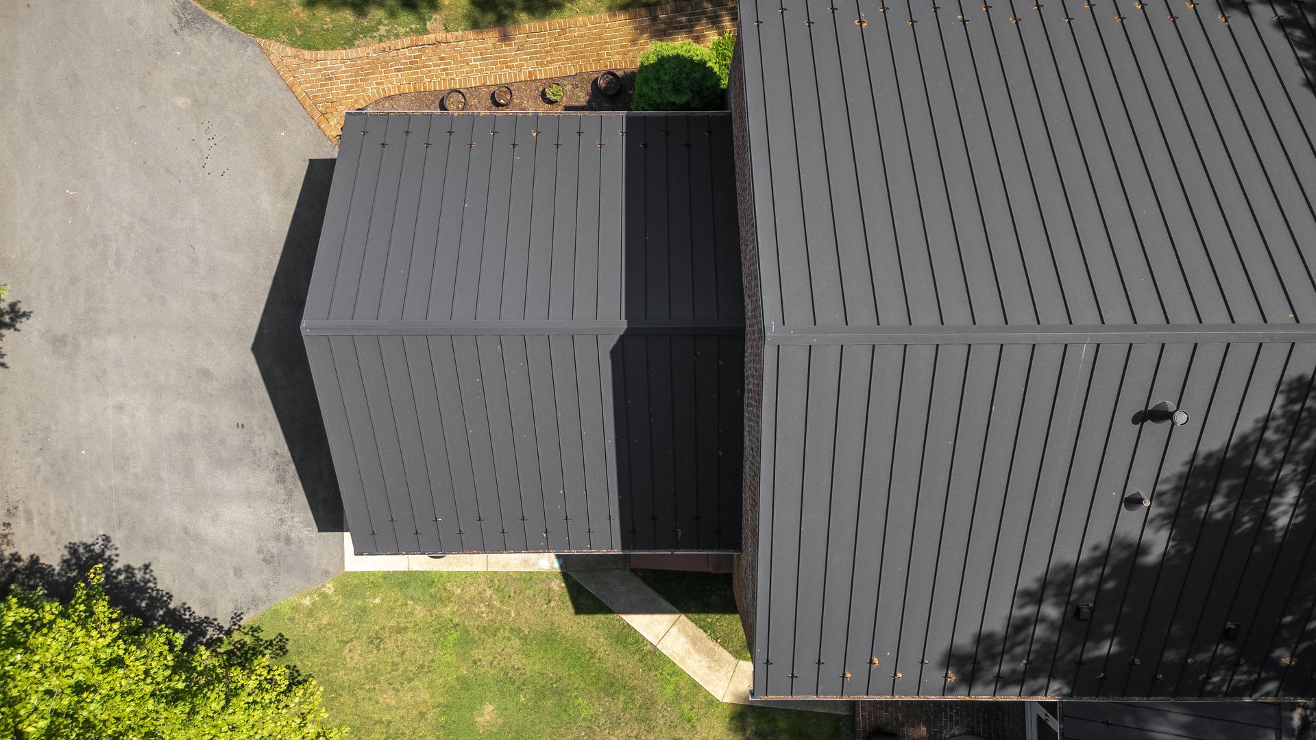 Overhead view of a dark gray house roof and driveway surrounded by green grass and trees.
