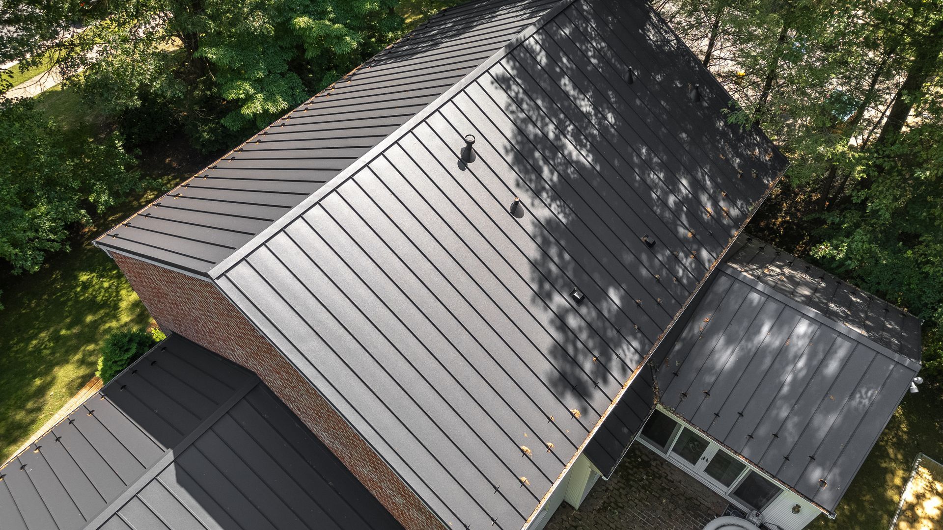 Overhead view of a house with a dark metal roof. The house is surrounded by green trees and grass.